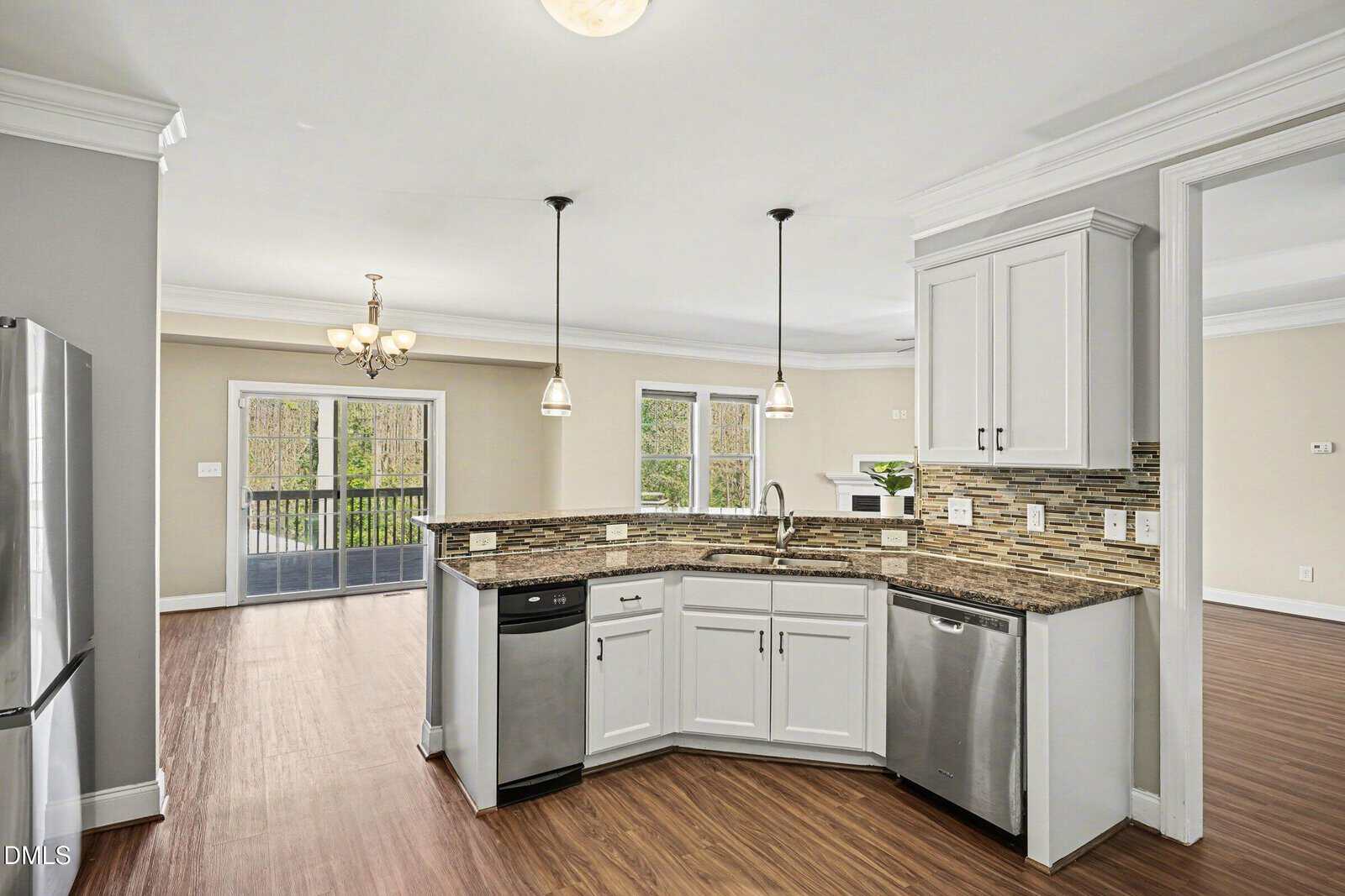 Modern kitchen with white cabinets, stainless steel appliances, mosaic tile backsplash, pendant lighting, and open view to dining area with large windows and wood floors.