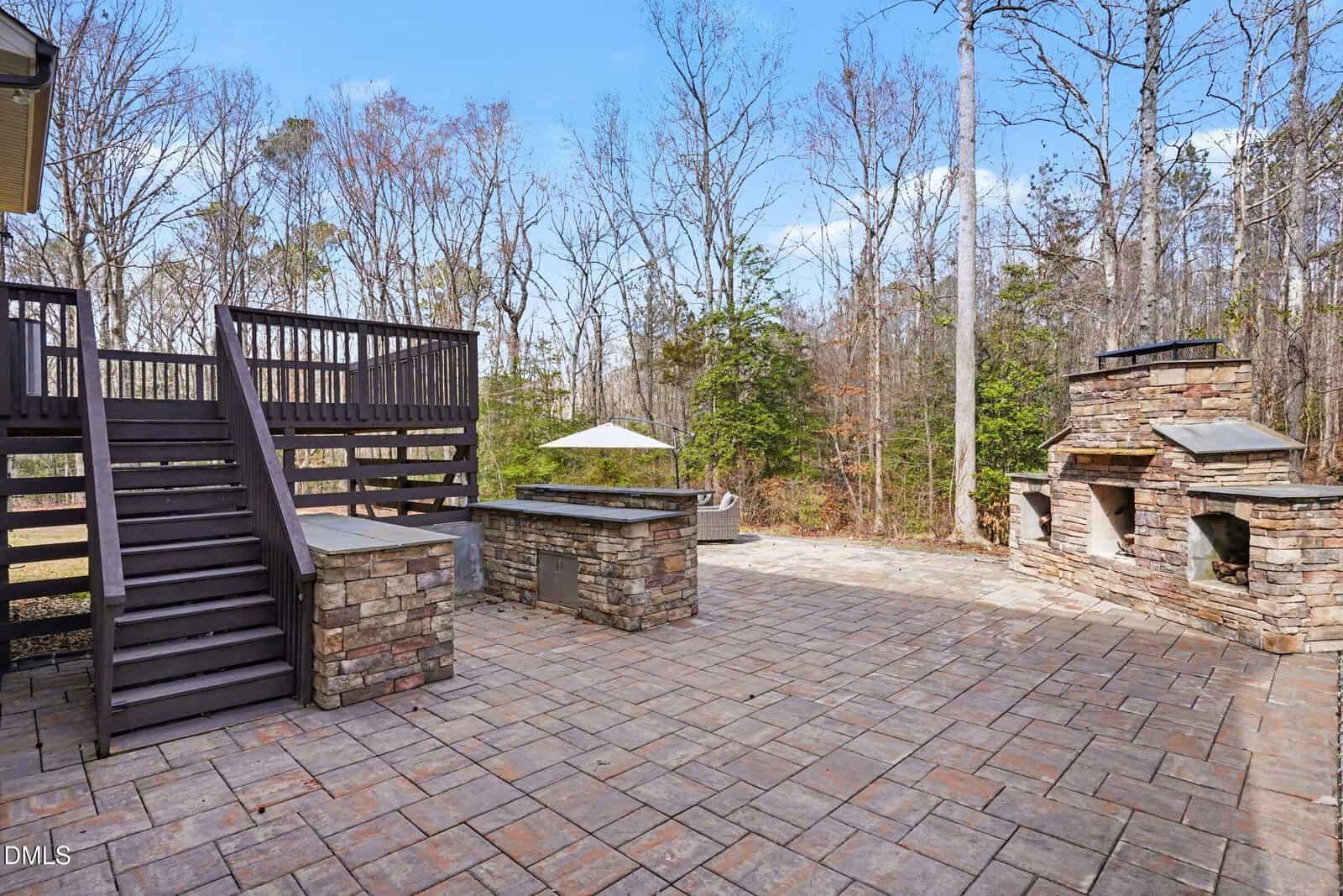 Spacious outdoor patio with stone flooring, built-in stone grill, fireplace, bar area, dark wooden staircase, and surrounded by trees and forest in the background.