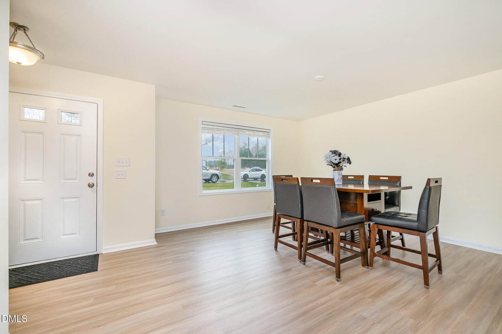 Bright dining area with a wooden table and six chairs, near a window and entrance door. Light wood flooring and cream walls create an open, inviting space. A vase with flowers sits on the table.