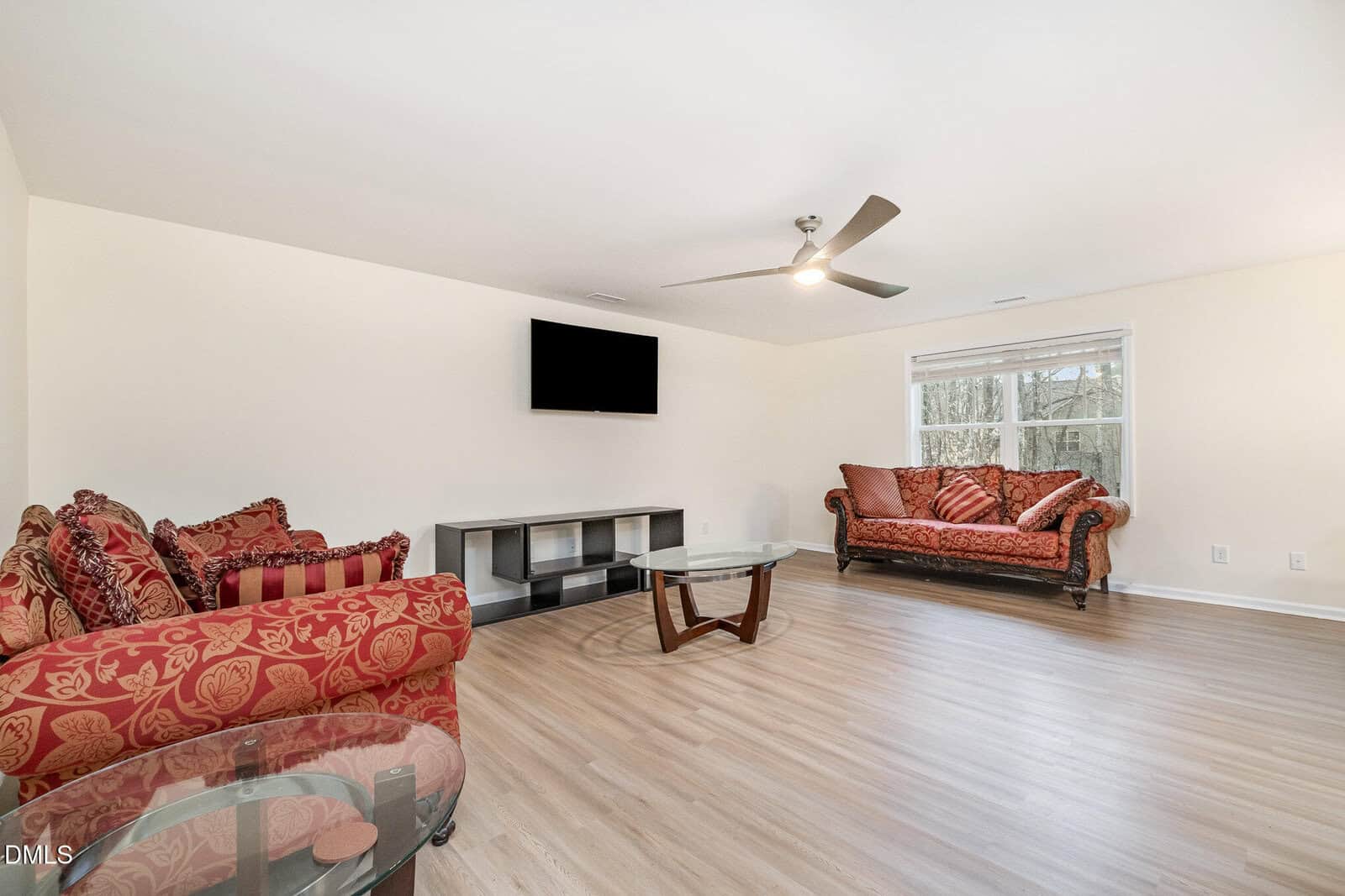 Spacious living room with two ornate red sofas, a glass coffee table, a wall-mounted TV, and large windows letting in natural light. The room has light wood floors and neutral walls.