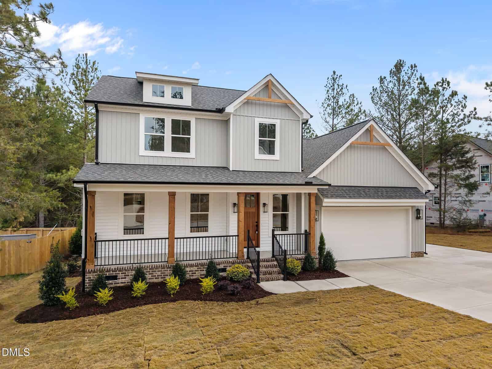 Two-story modern farmhouse with light gray siding, dark roof, wooden porch posts, front steps, attached two-car garage, landscaped yard, and driveway, set against a backdrop of trees and blue sky.
