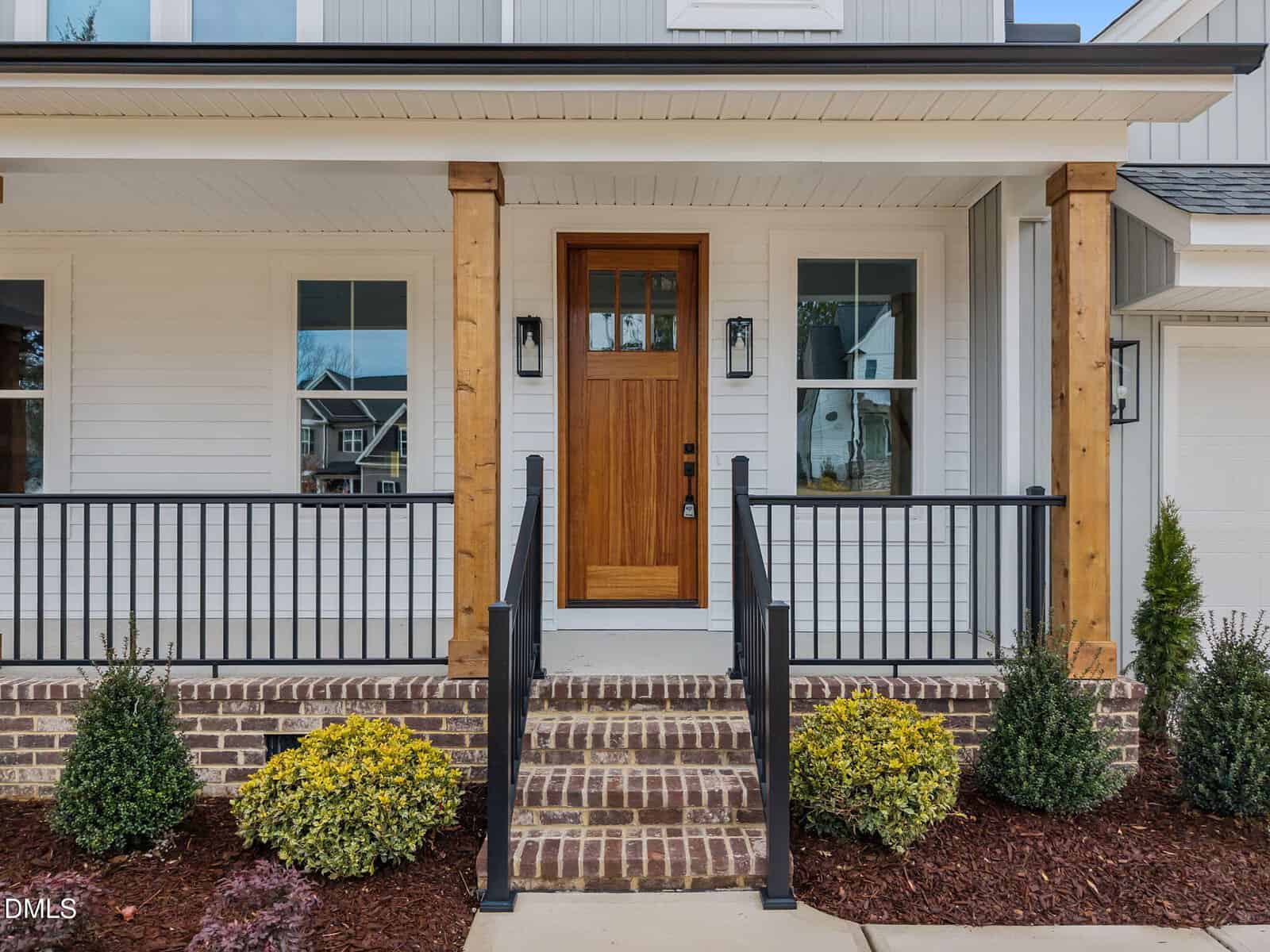 A modern house entrance with a wooden front door, black railing, steps with brick risers, and landscaped shrubs along the walkway. White siding and two wooden columns frame the porch.