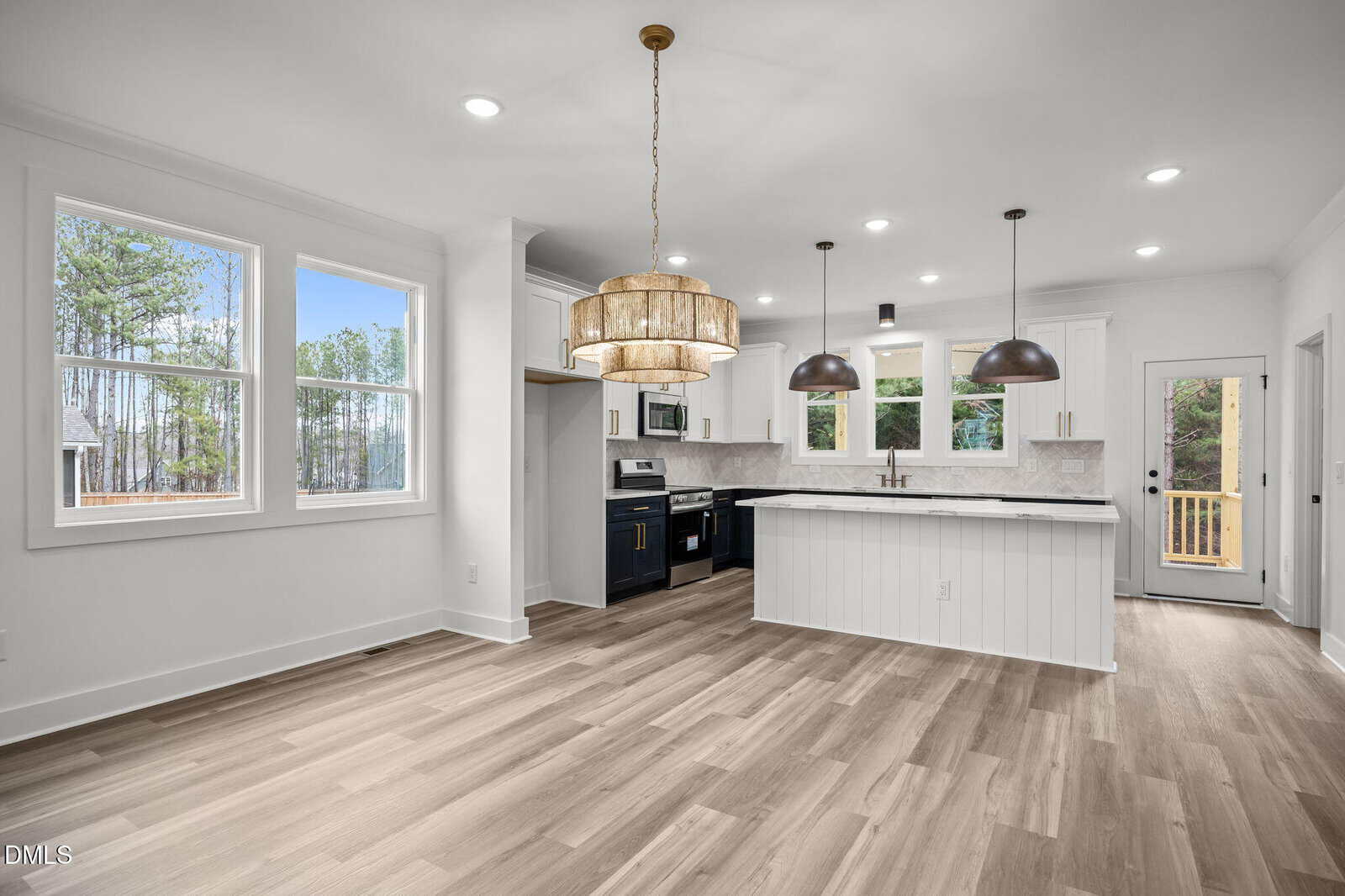 Modern kitchen and dining area with light wood flooring, white walls, large windows, and pendant lights above an island. Stainless steel appliances and a door leading outside are visible. The space is bright and open.