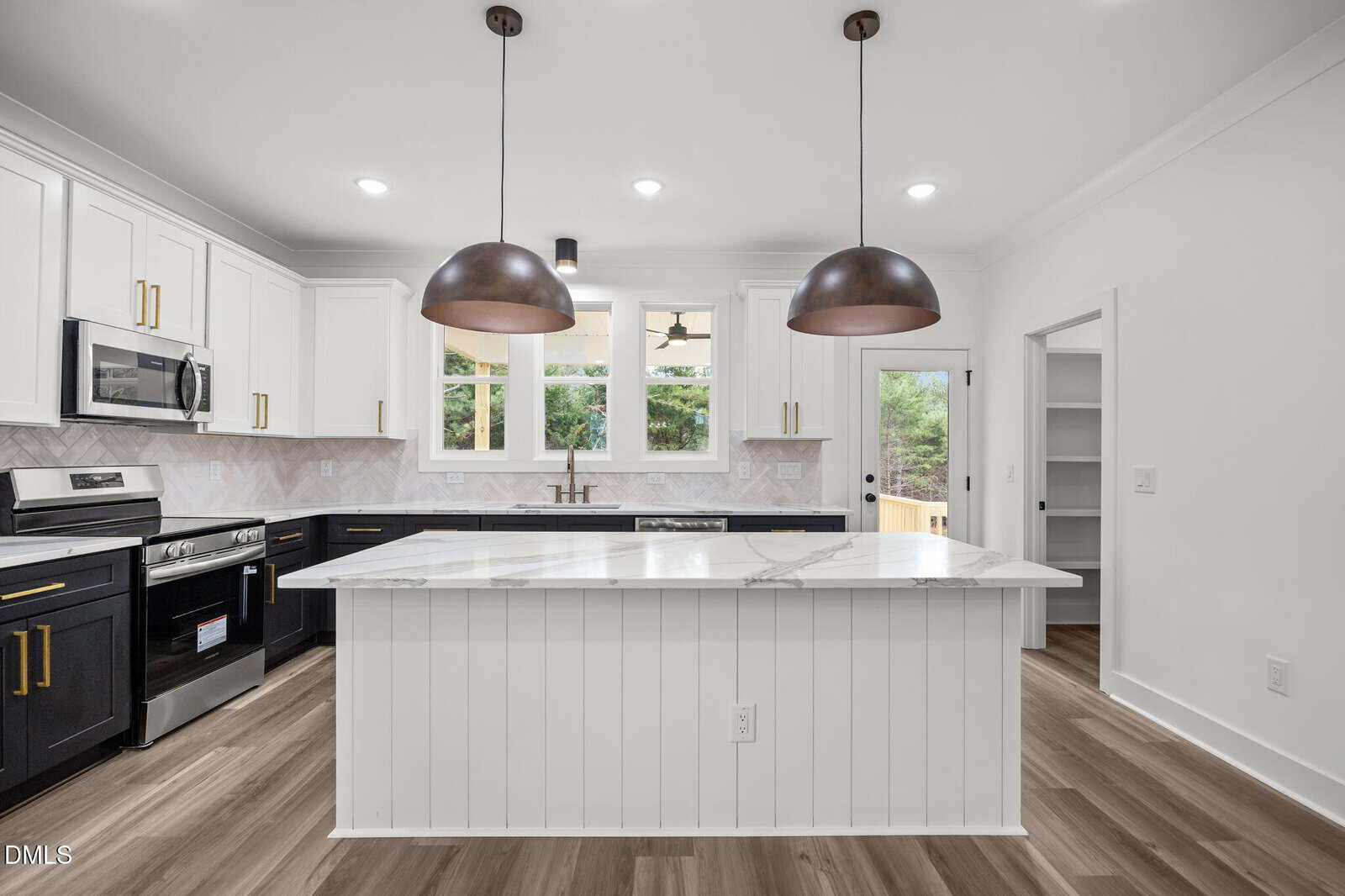 Modern kitchen with black and white cabinetry, stainless steel appliances, marble island, two large pendant lights, wood-style flooring, and a window above the sink providing natural light. Doorway leads to a pantry.