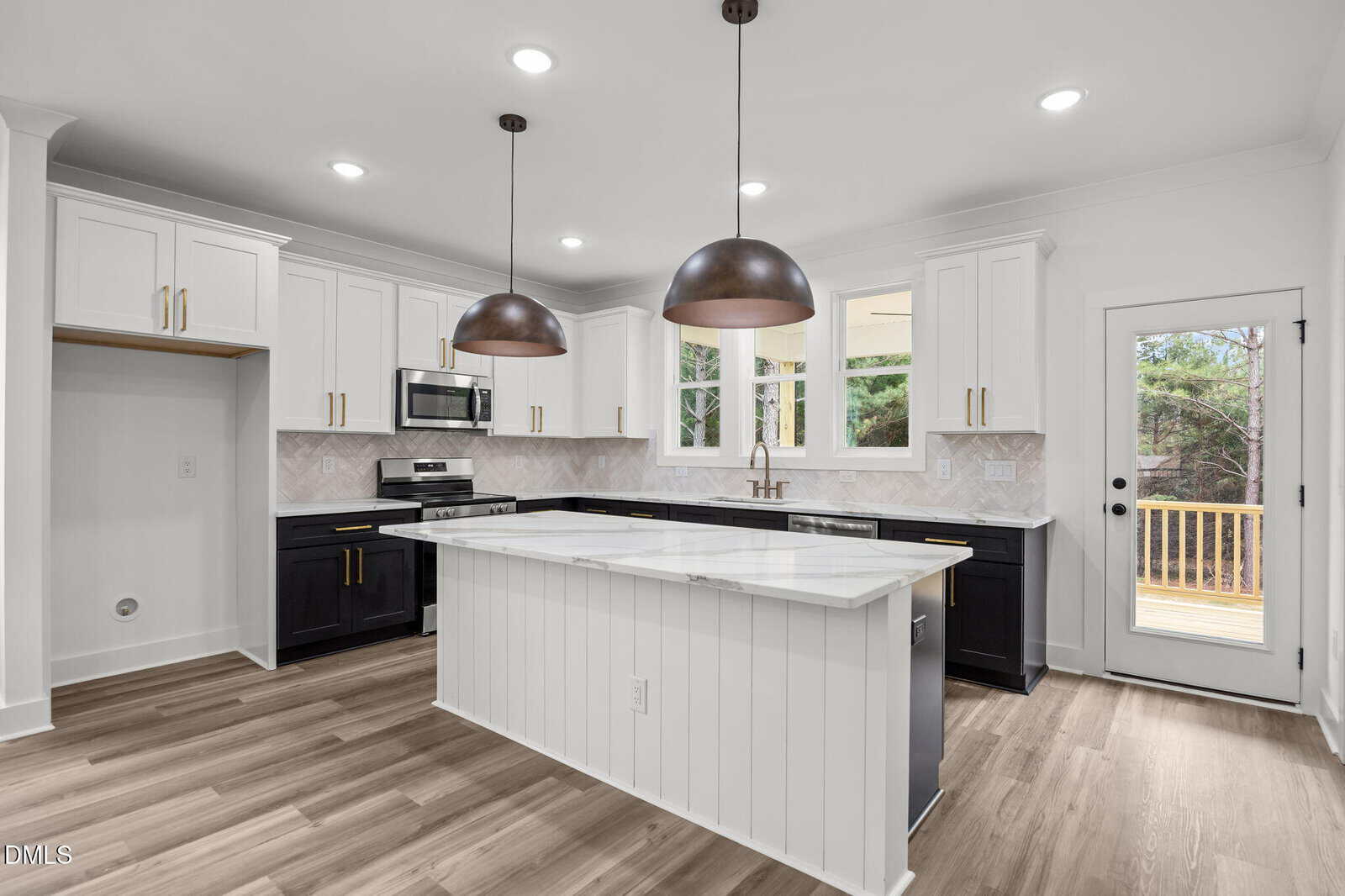 Modern kitchen with two-tone cabinets, white upper and black lower, a white island with marble countertop, pendant lights, stainless steel appliances, wood floors, and a door leading to a deck outside.