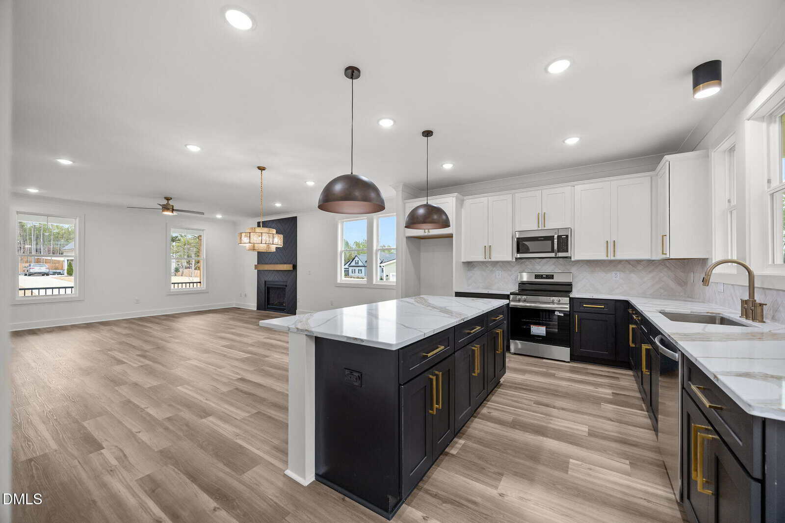 Modern kitchen with black and white cabinets, marble countertops, stainless steel appliances, pendant lights, and open layout connected to a living area with large windows, wood flooring, and a fireplace.