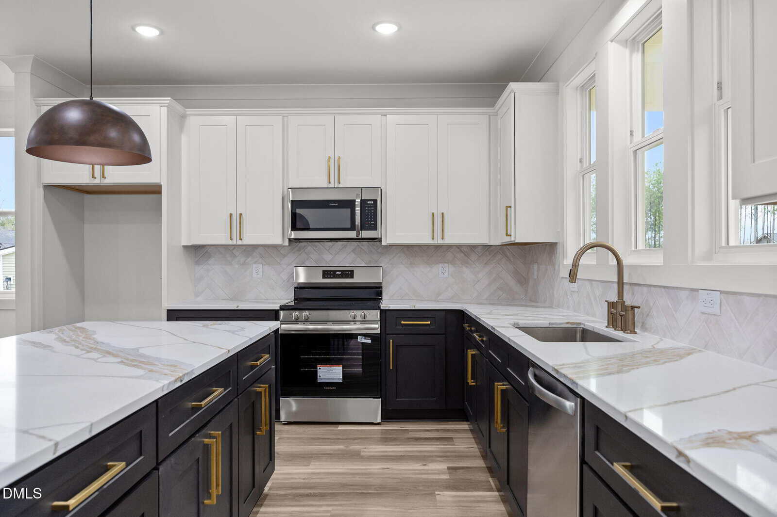 Modern kitchen with white upper cabinets, dark lower cabinets, gold handles, marble countertops, stainless steel appliances, a large sink under windows, and a pendant light hanging from the ceiling.