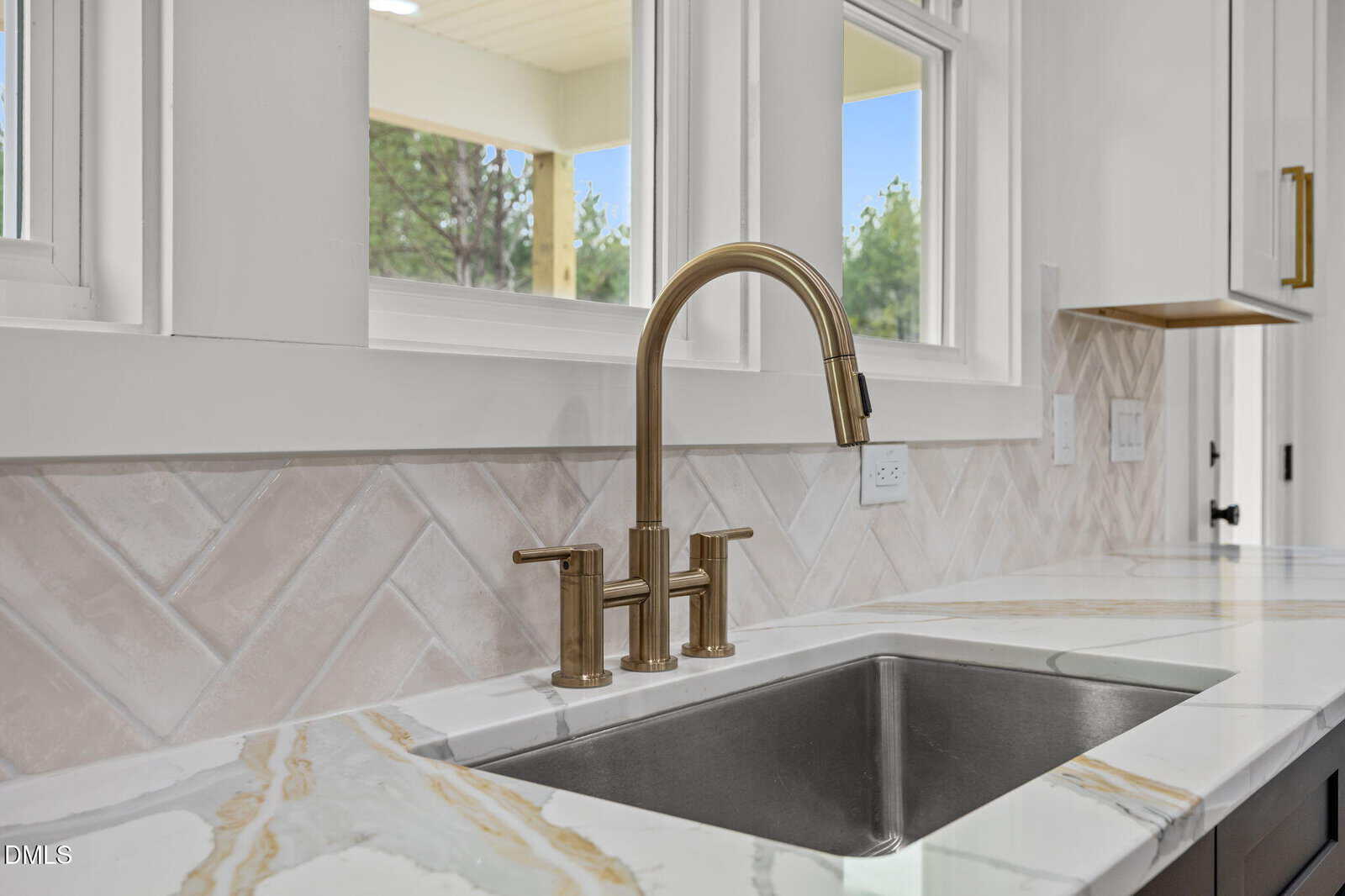 Modern kitchen sink with a gold faucet, marble countertop, and light herringbone tile backsplash. Two windows above the sink allow natural light into the space.