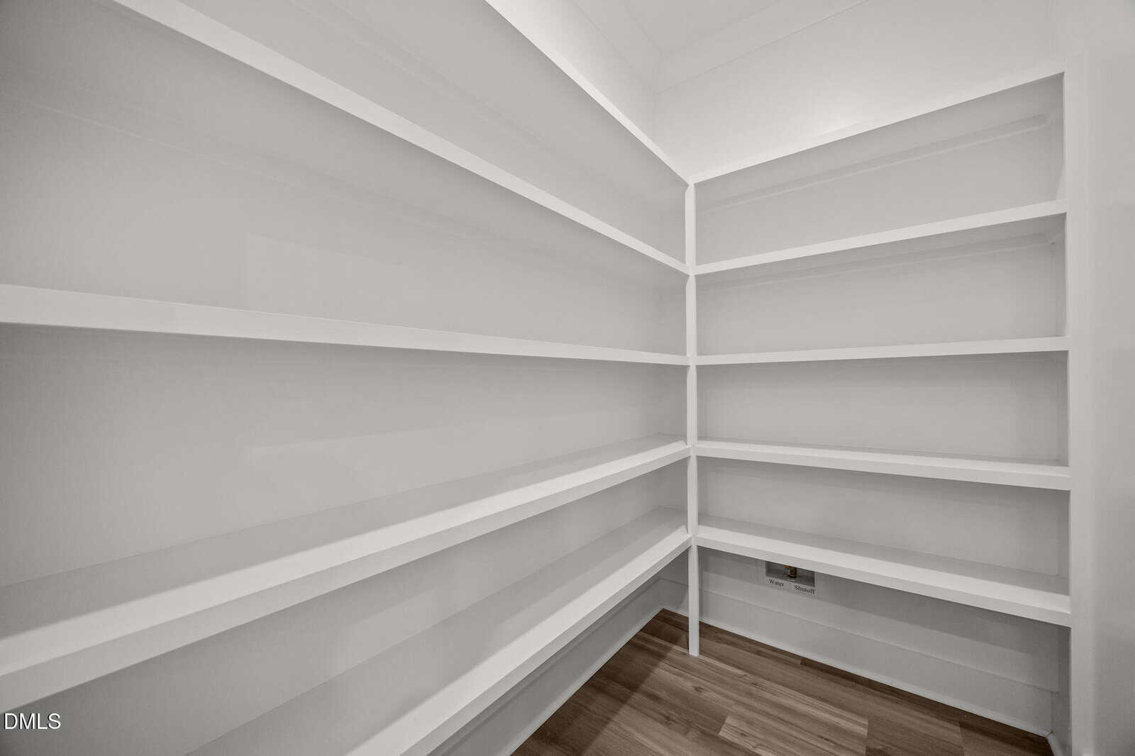 A spacious, empty walk-in pantry with white built-in shelves along two walls and a light wood floor. The room is brightly lit and clean, with modern, minimalist design.