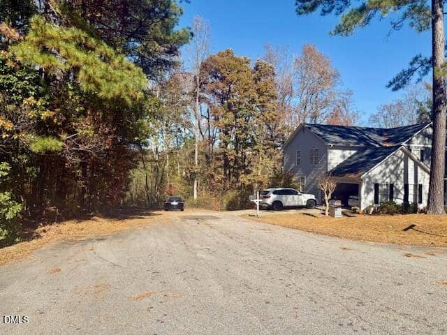 A quiet suburban street bordered by trees with two parked vehicles in the driveway of a two-story house on the right side. Fallen leaves are scattered across the road and lawns. The sky is clear and blue.