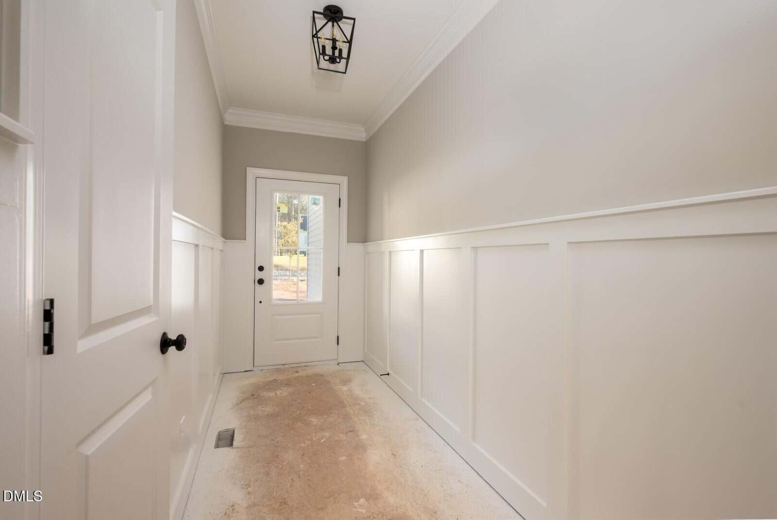 A narrow hallway with beige walls and white wainscoting leads to a closed white door with a window. The floor appears unfinished, and a black light fixture hangs from the ceiling.