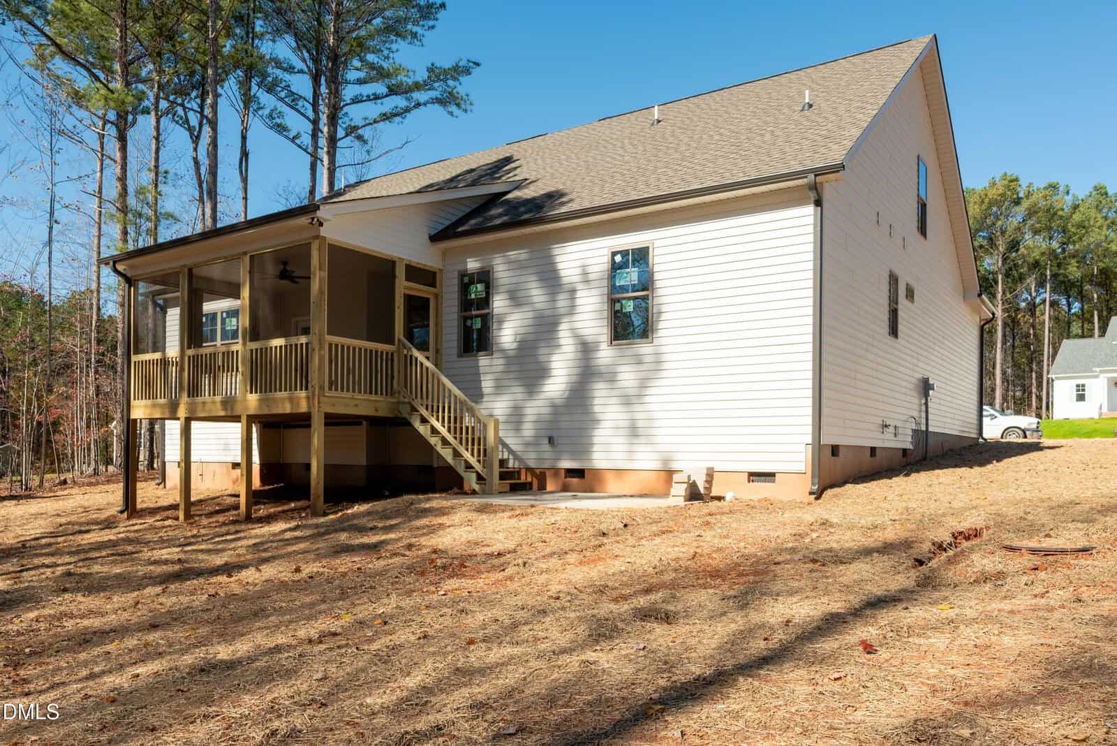 A single-story white house with gray roof, screened porch, wooden steps, and multiple windows, set on a dirt lot with scattered trees and a smaller house in the background.