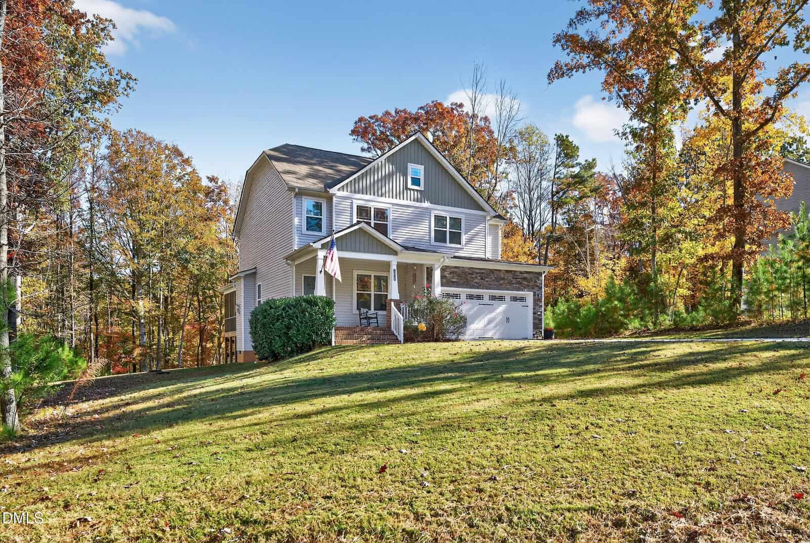 A two-story house at 1318 Sourwood Drive in Wake Forest, with gray siding and white trim, features a covered front porch, an American flag, and a two-car garage, set on a grassy lawn among autumn trees under a blue sky.