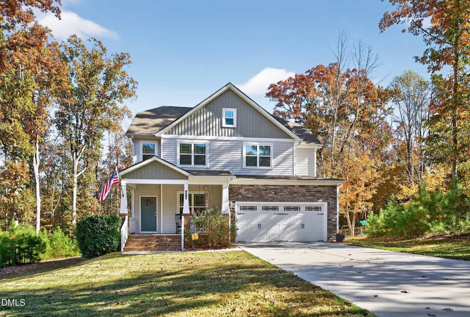 A two-story house with gray siding, stone accents, and a covered front porch. An American flag is displayed near the entrance. A driveway leads to a double garage. Trees with autumn foliage surround the property.