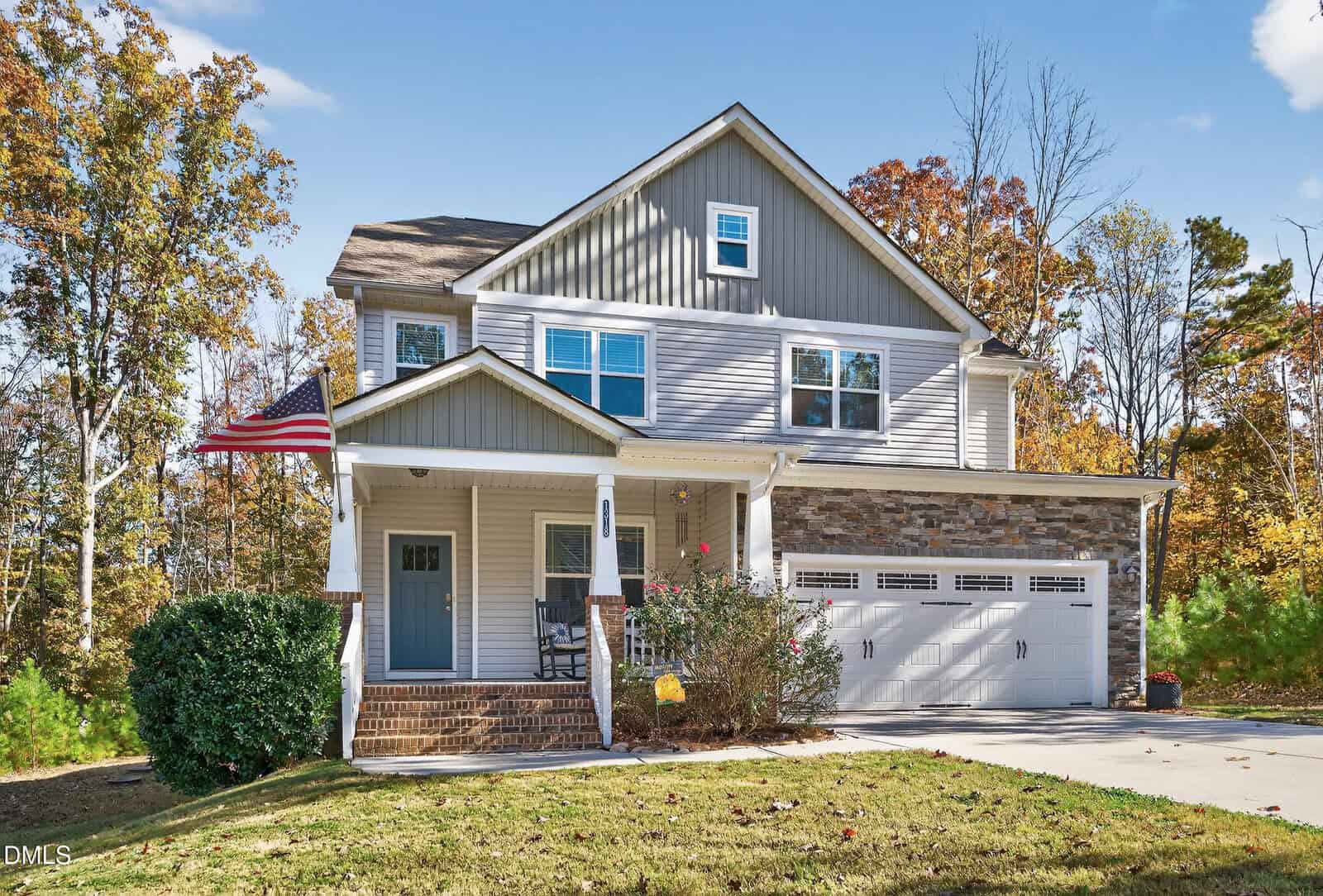 Two-story suburban house with gray siding, stone accents, and a covered front porch. An American flag is displayed by the entrance. There is a double garage and a concrete driveway. Trees surround the property.