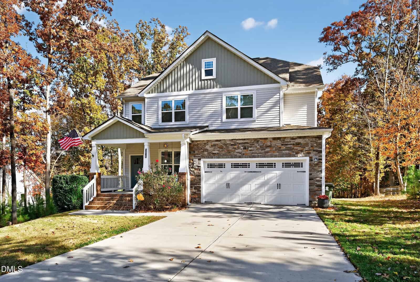 A two-story suburban house with stone and siding exterior, a front porch with an American flag, and a double garage. Trees with autumn foliage surround the property, and a concrete driveway leads to the garage.