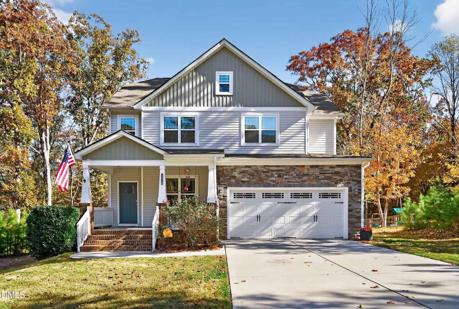 A two-story suburban house with gray siding, stone accents, and a covered front porch. An American flag hangs by the entrance. The house has a double garage and is surrounded by trees with autumn foliage.