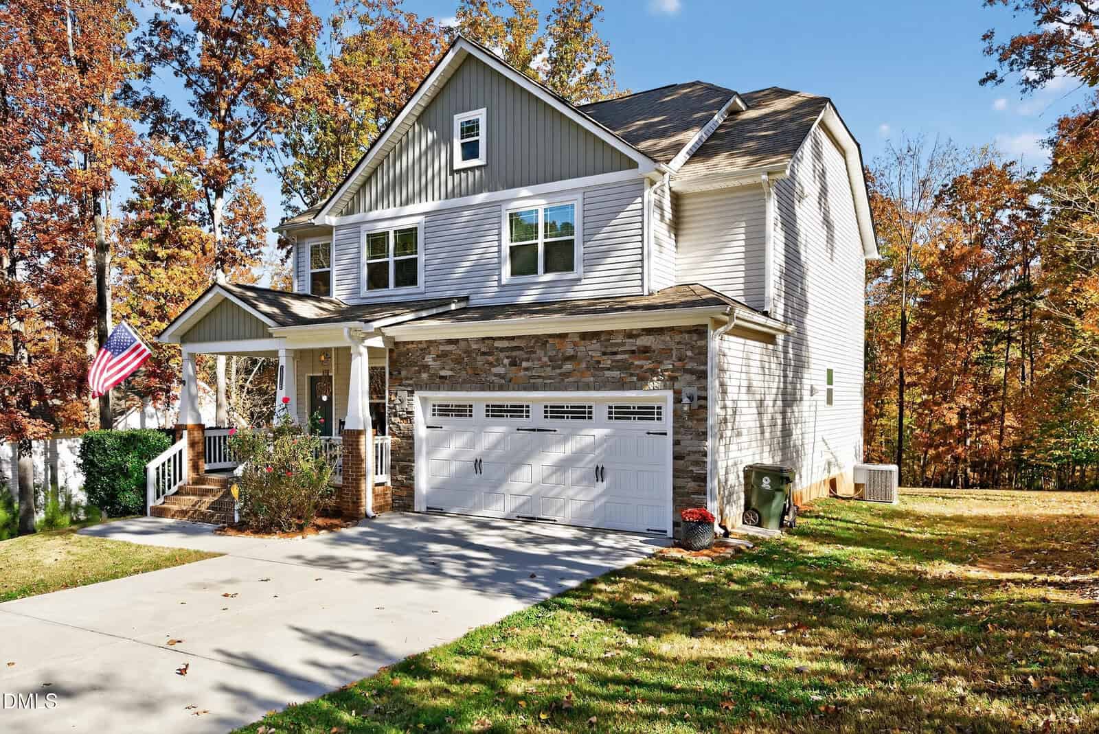 Two-story suburban house with stone and gray siding exterior, a white garage door, and a covered front porch with white railings. An American flag hangs by the entrance. The yard is grassy with trees in the background.