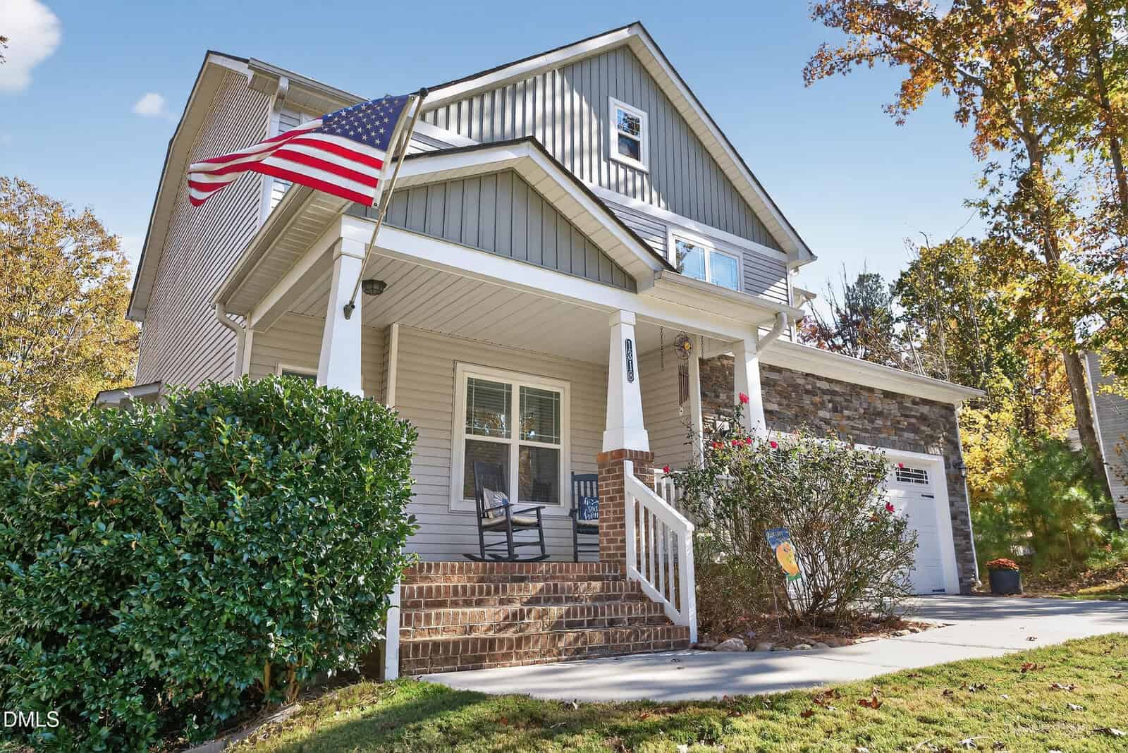 Two-story suburban house with a covered front porch, American flag, brick steps, and stone accents. A driveway leads to a two-car garage. Green shrubs and trees surround the house under a clear blue sky.