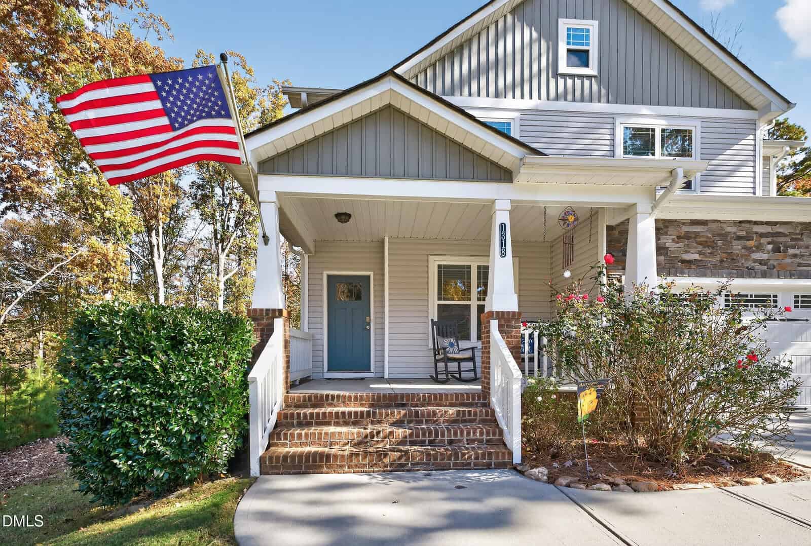 A two-story suburban house with a covered front porch, brick steps, a rocking chair, an American flag, and bushes in front. The exterior is gray with white trim and stone accents.