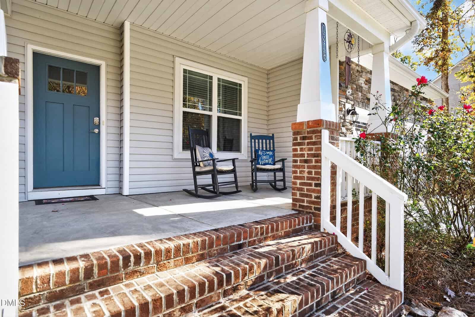 A front porch with brick steps, two black rocking chairs, a small side table, and a light blue door. White railings and beige siding are visible, with some flowering bushes by the stairs.
