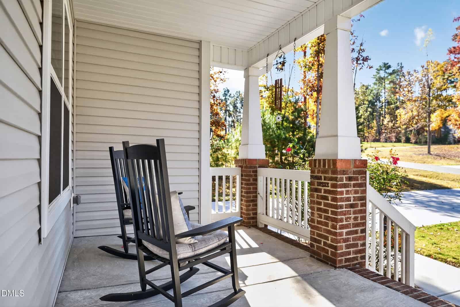 A front porch with two black rocking chairs, white railings with brick columns, and a concrete floor. Trees with autumn foliage and a sidewalk are visible in the background.