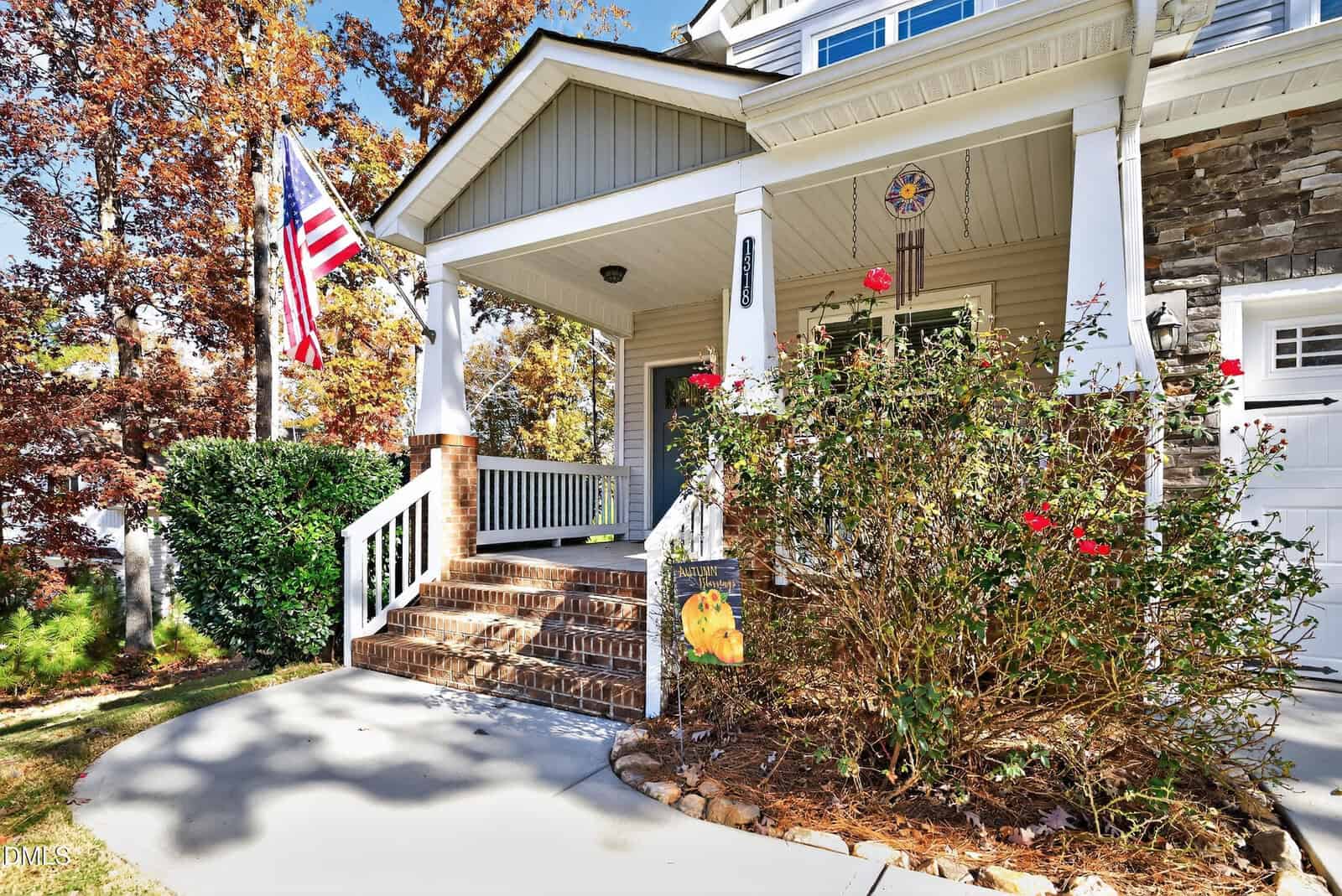 A house front with stairs leading to a porch, an American flag, blooming rose bushes, a butterfly flag, brick and stone exterior, and autumn trees in the background.