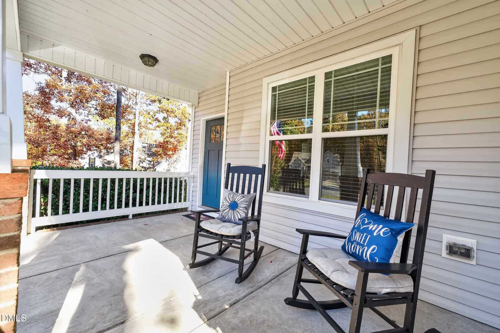 A cozy front porch with two black rocking chairs, one with a “Home Sweet Home” pillow, sits beside a window and a blue front door. Trees with autumn leaves are visible in the background.