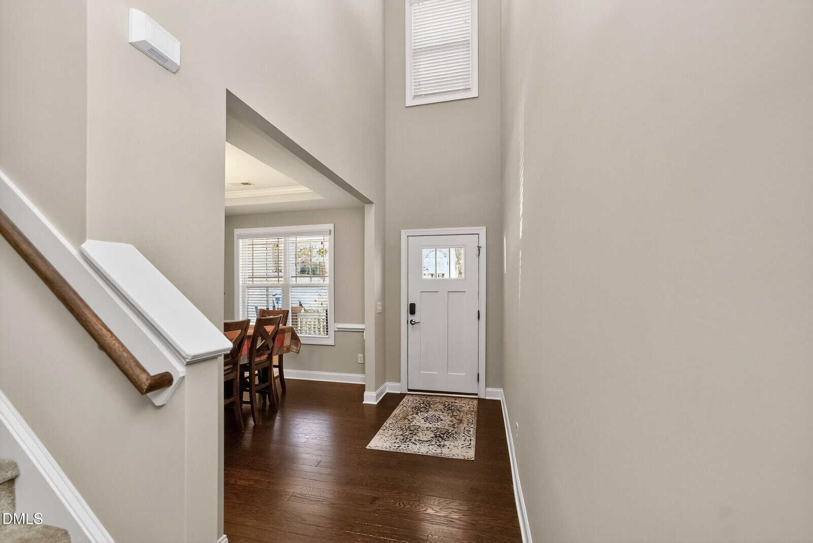 Entryway with a white front door, tall ceiling, and window above. Dark wood floors, a small rug, and stairs with a white banister are visible, with a dining area to the left. Bright, neutral-colored walls.