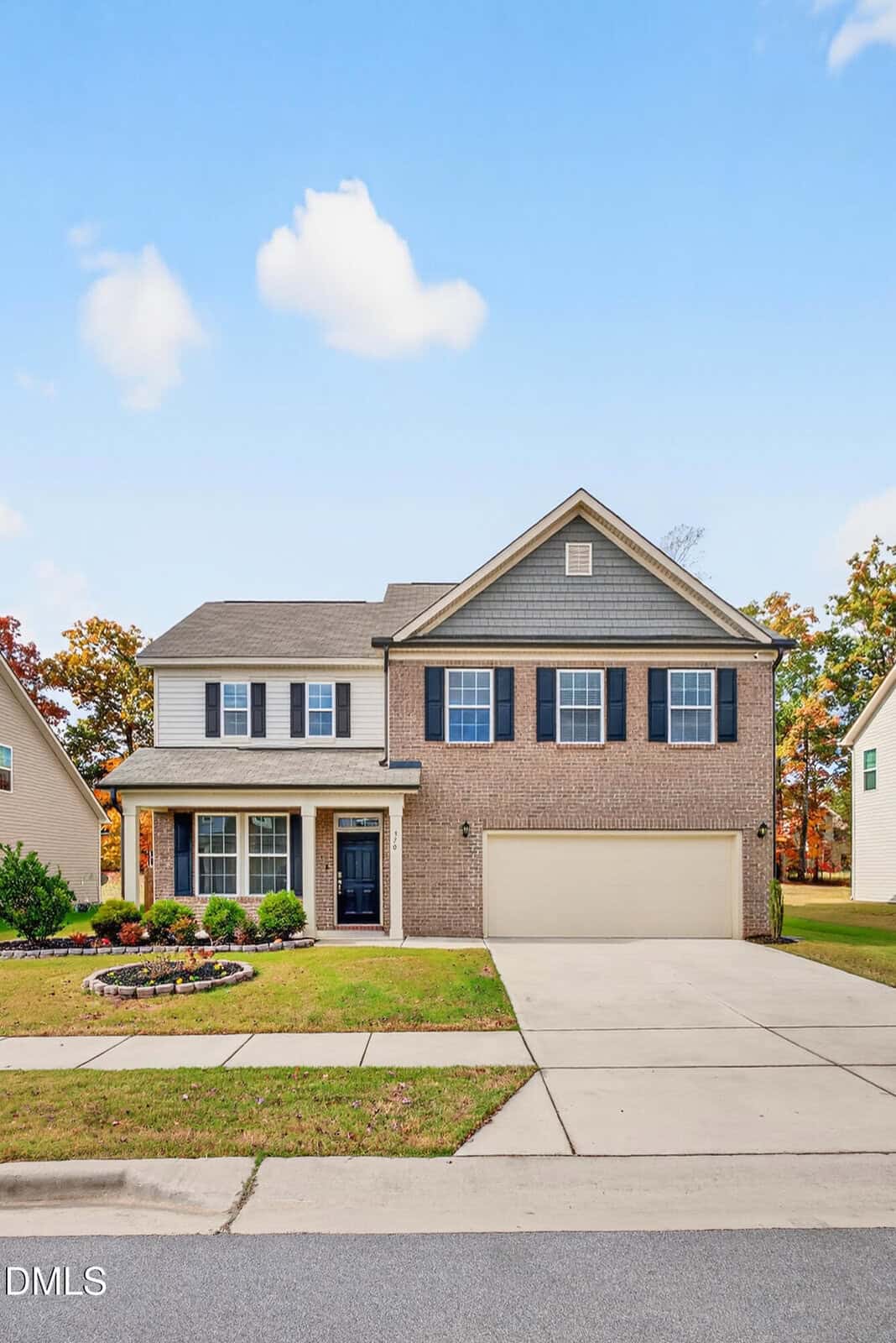 Two-story suburban house with a brick and siding exterior, blue shutters, a two-car garage, and a landscaped front yard, set against a clear blue sky with a few clouds.