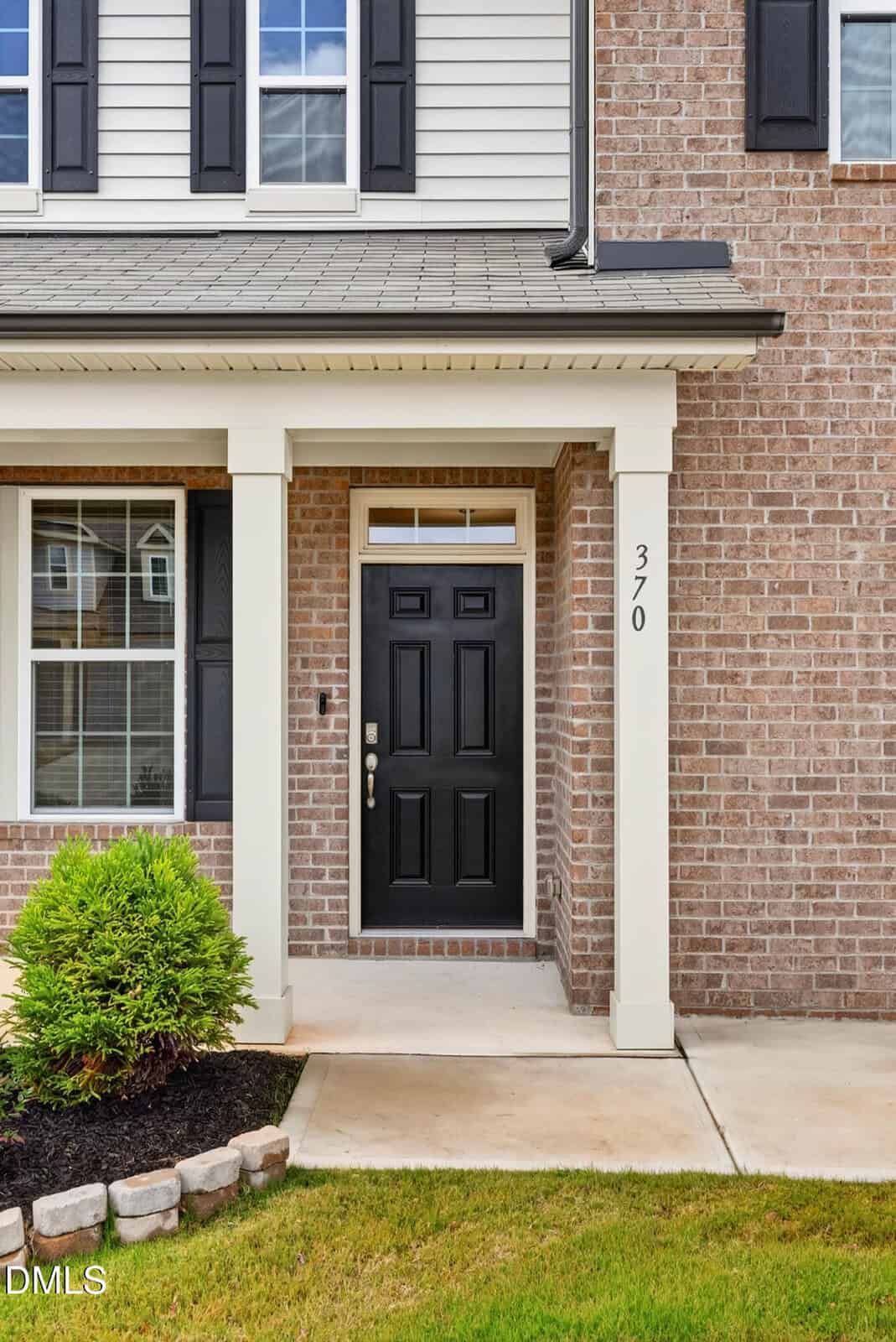 A front entryway of a brick house featuring a black door with a glass transom window above, white trim, the number 370 on a column, and a small shrub near the walkway.