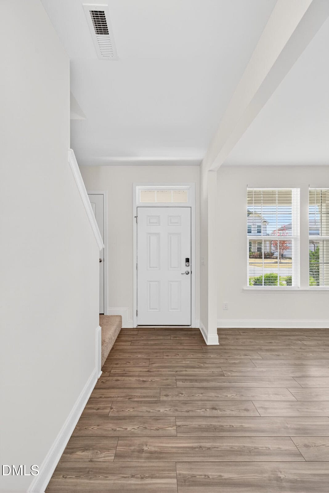 A bright entryway with white walls, a white front door, wood-style flooring, stairs on the left, and large windows on the right letting in natural light.