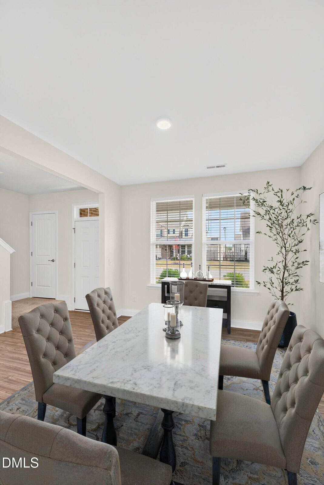 A modern dining room with a rectangular marble table, six tufted chairs, a rug, large windows with blinds, a potted plant, and light gray walls. Natural light fills the space.