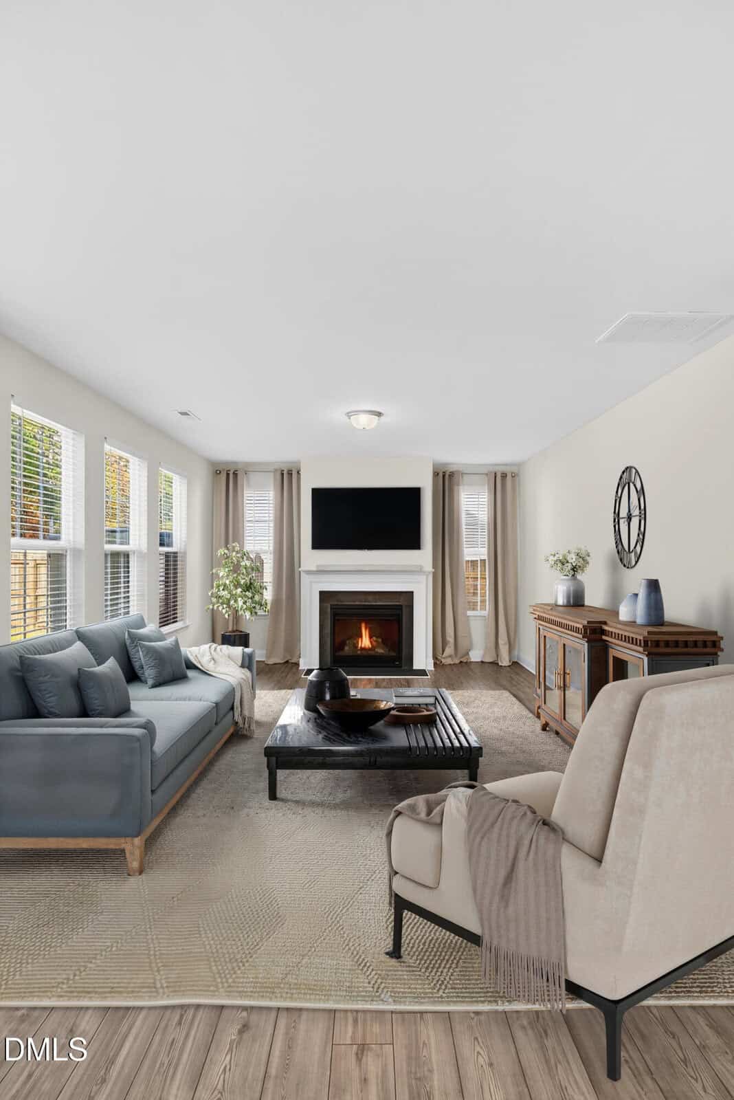 A modern living room with a blue sofa, beige armchair, coffee table, and fireplace. Large windows let in natural light. Neutral decor includes plants and a sideboard with vases, all on a light patterned rug.