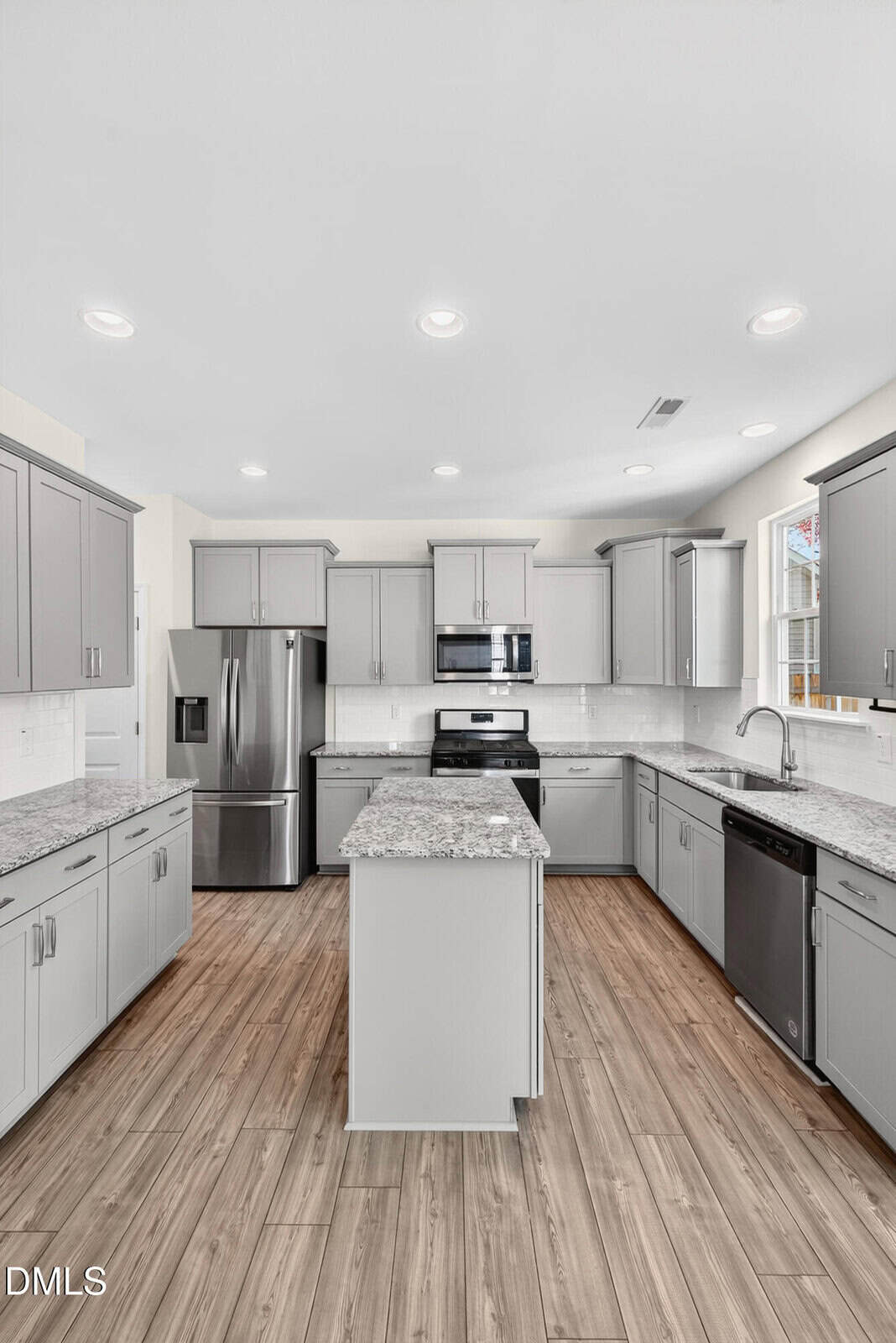 Modern kitchen with gray cabinets, stainless steel appliances, a central island with a granite countertop, white subway tile backsplash, and wood-look flooring under bright ceiling lights.