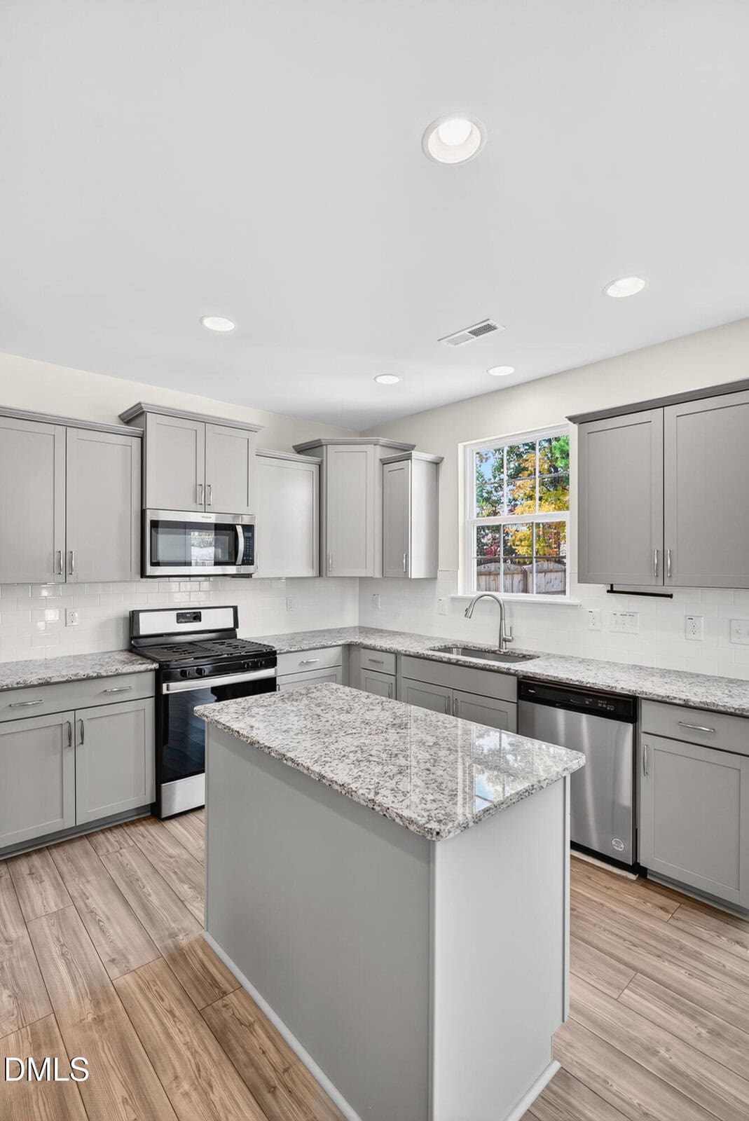 Modern kitchen with gray cabinets, stainless steel appliances, granite countertops, white subway tile backsplash, light wood flooring, and a central island. A window above the sink lets in natural light.