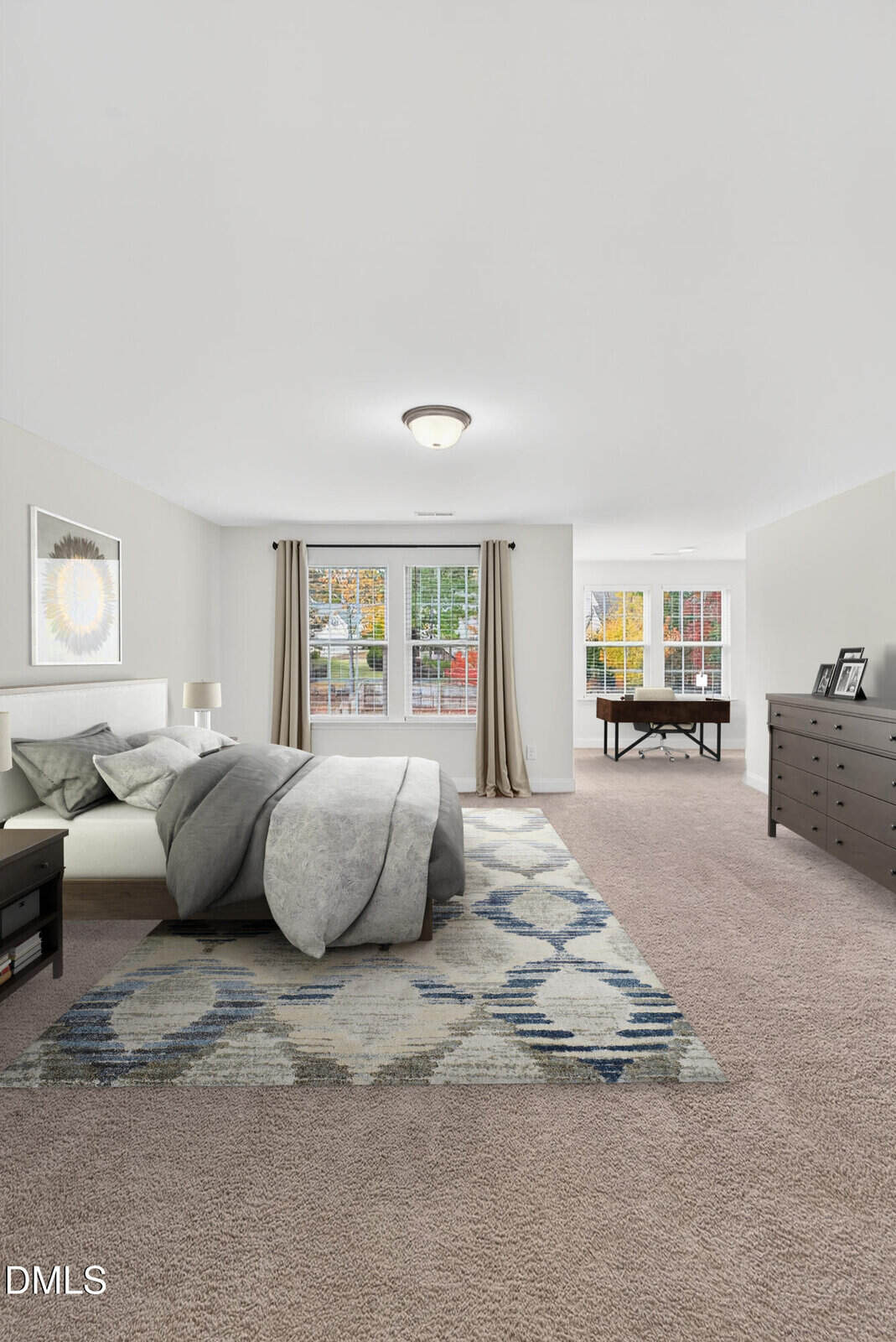 Spacious bedroom with a large bed, gray bedding, geometric rug, nightstands, dresser, and a desk near bright windows with view of trees. Room features neutral tones and carpeted floor.