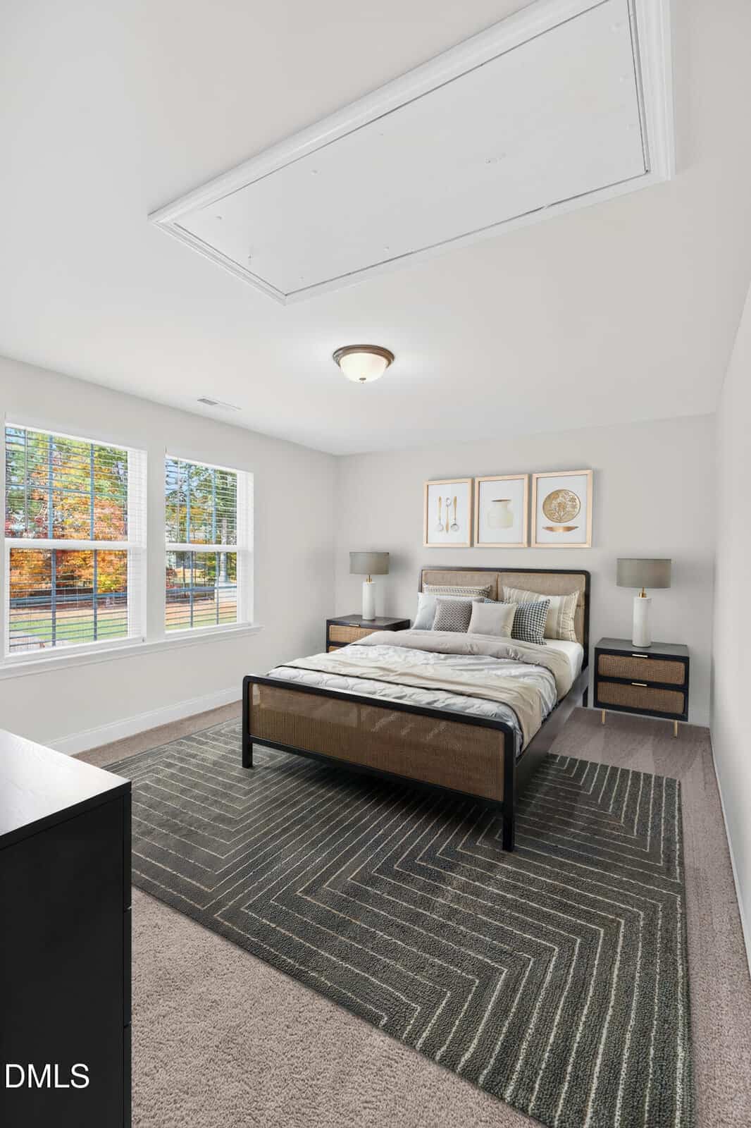 A bright bedroom with large windows, a double bed with striped bedding, two bedside tables with lamps, neutral walls, and a geometric patterned area rug on carpeted flooring. Trees are visible outside.