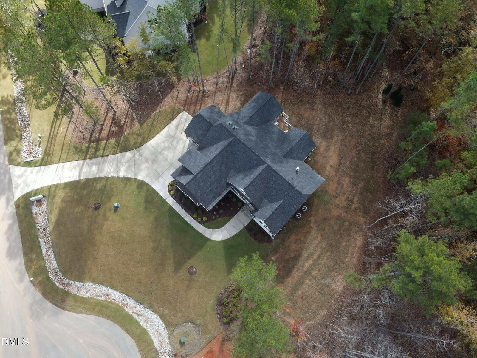 Aerial view of a single-story house with a dark roof, surrounded by trees and a lawn. A curved driveway leads to the house from a cul-de-sac, and the backyard borders a wooded area.