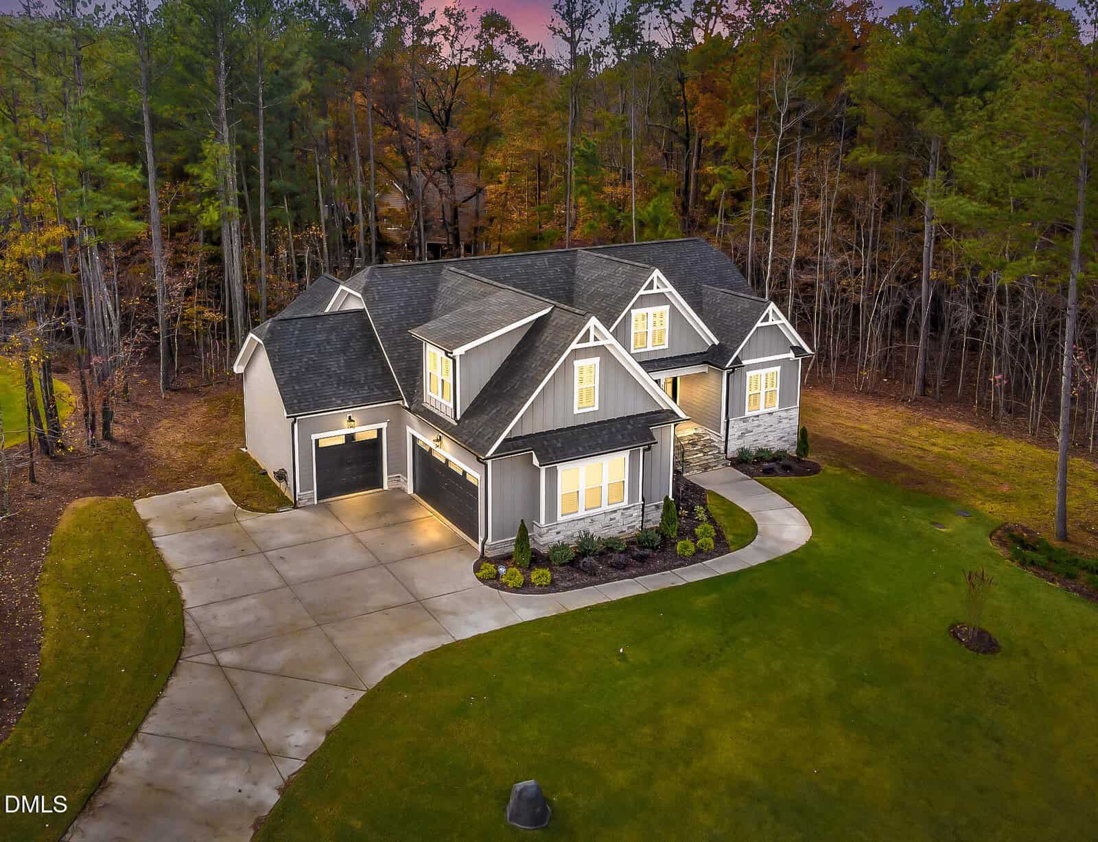 A gray, two-story suburban house with a three-car garage is surrounded by a green lawn and bordered by tall trees. The driveway curves toward the house, and exterior lights are on at dusk.