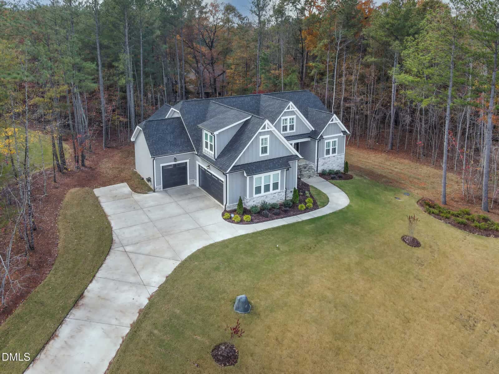 Aerial view of a two-story gray house with a dark roof, attached garage, and curved driveway, surrounded by grass and bordered by trees in the background.