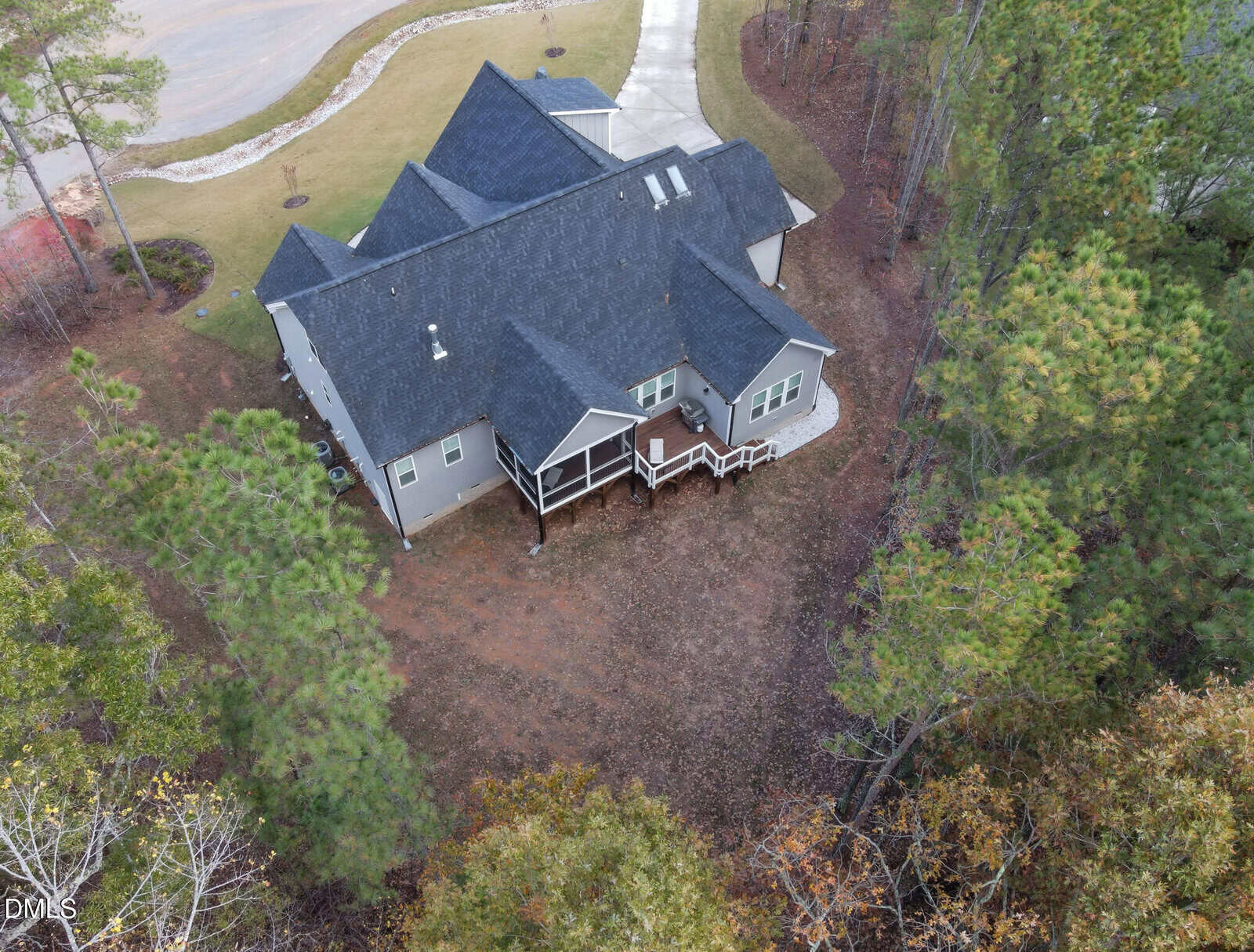 Aerial view of a two-story house with a dark gray roof, covered front porch, and driveway. The house is surrounded by trees and has a cleared yard with patches of grass and dirt.