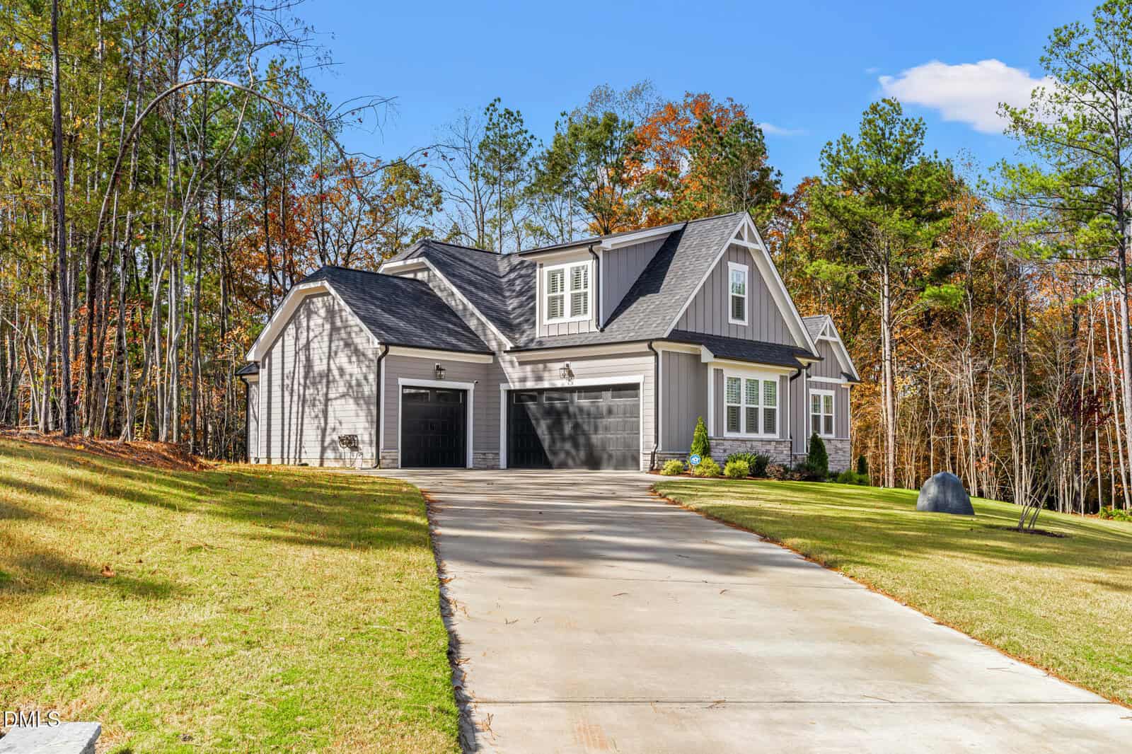 A gray two-story house with a double garage and dormer windows sits at the end of a long concrete driveway, surrounded by a grassy lawn and trees with autumn foliage.