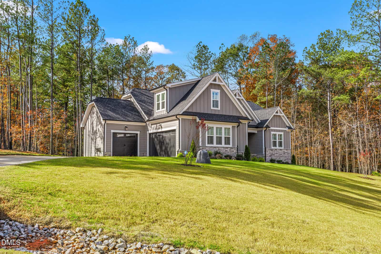 Single-story gray house with a gabled roof, attached garage, and stone accents, set on a sloped grassy lawn with trees in the background under a blue sky.