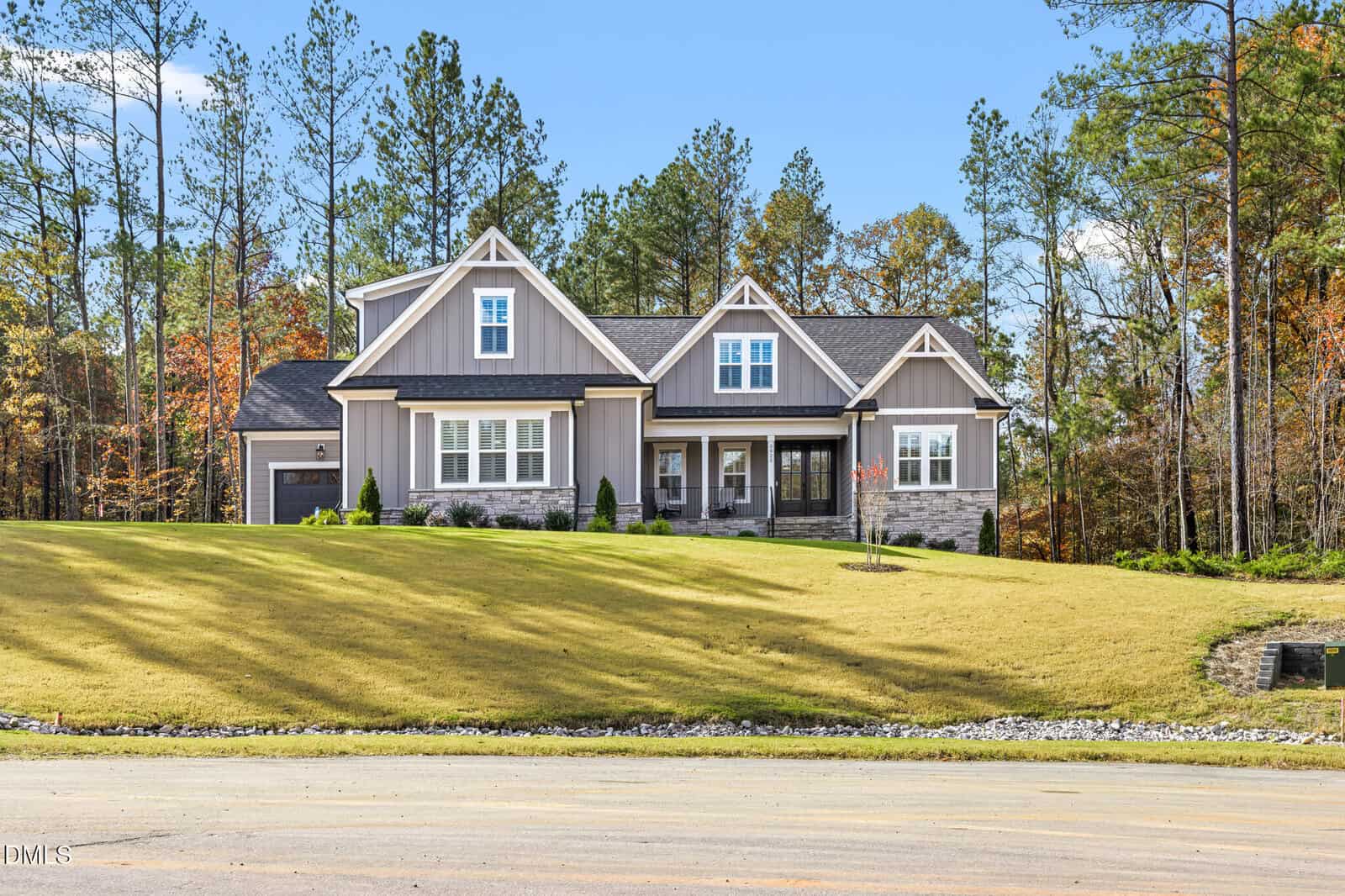 A modern single-story house with gray siding and stone accents sits on a large, sloped lawn with young landscaping, surrounded by tall trees under a clear blue sky.