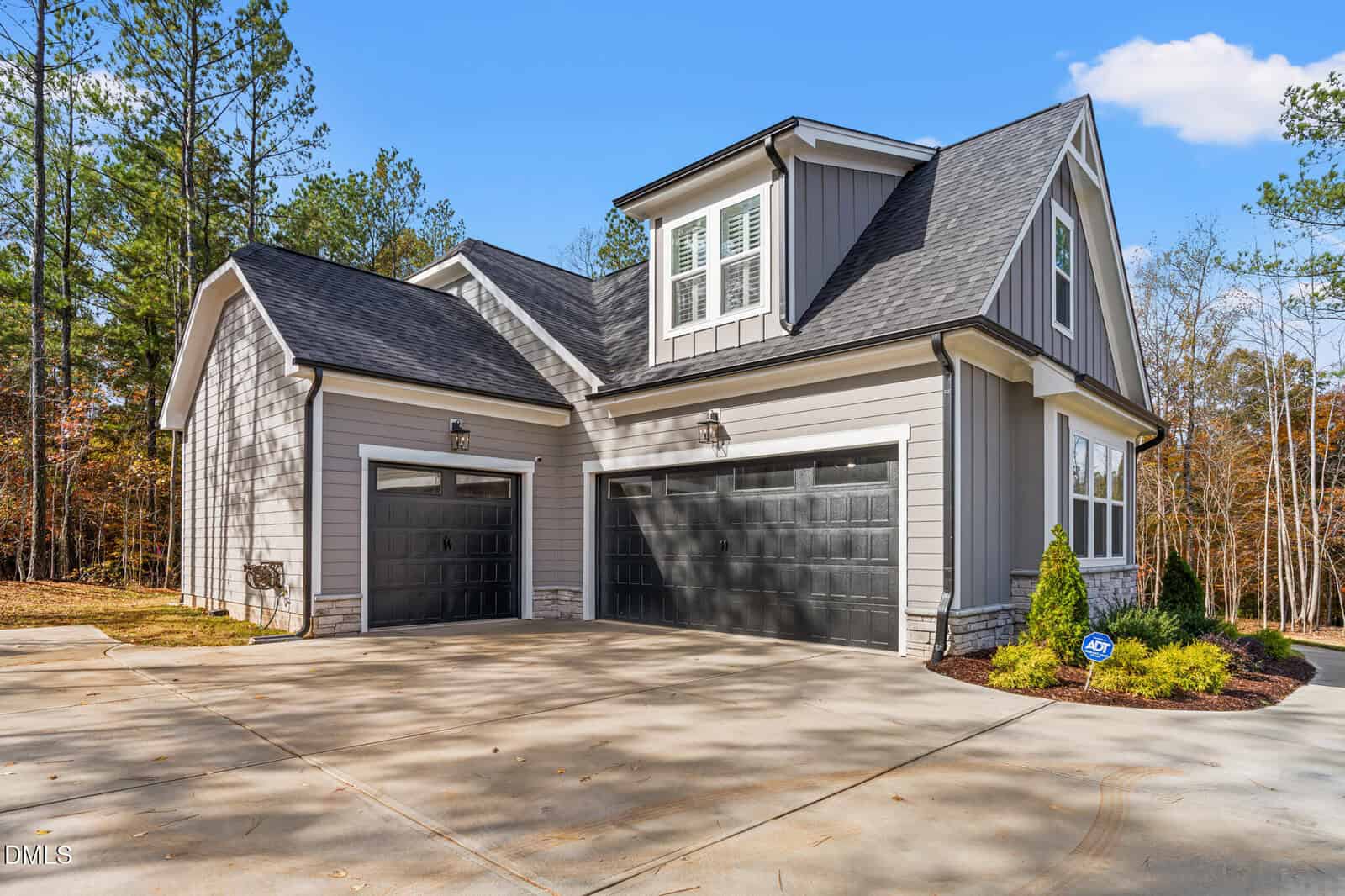 Modern, two-story gray house with black double garage doors, white trim, and a gabled roof. Surrounded by trees, with a concrete driveway and well-kept landscaping under a clear blue sky.