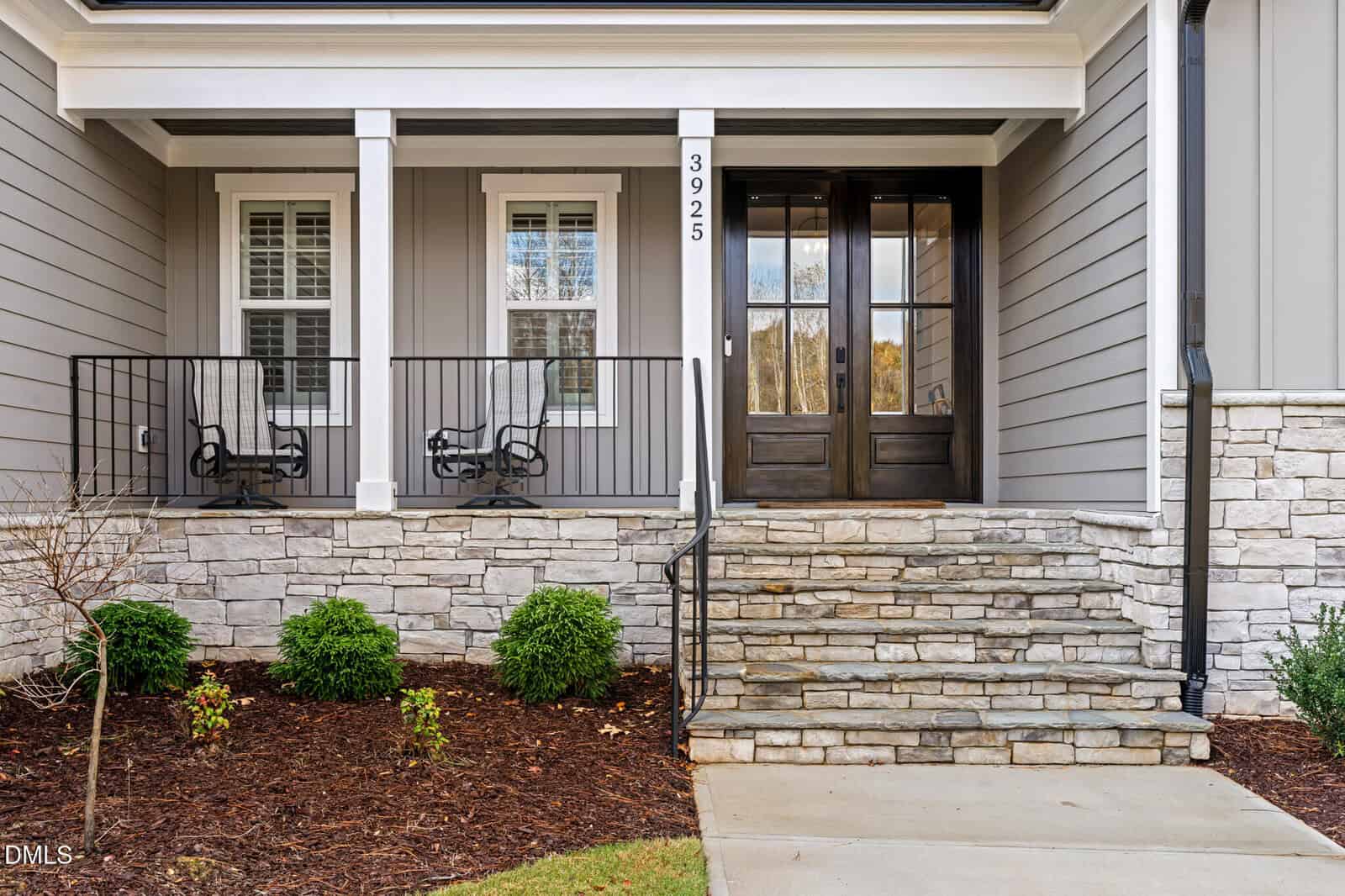 Front entrance of a house with stone steps, double wooden doors, two windows, a small porch with black railings and rocking chairs, gray siding, and landscaped bushes in mulched beds.
