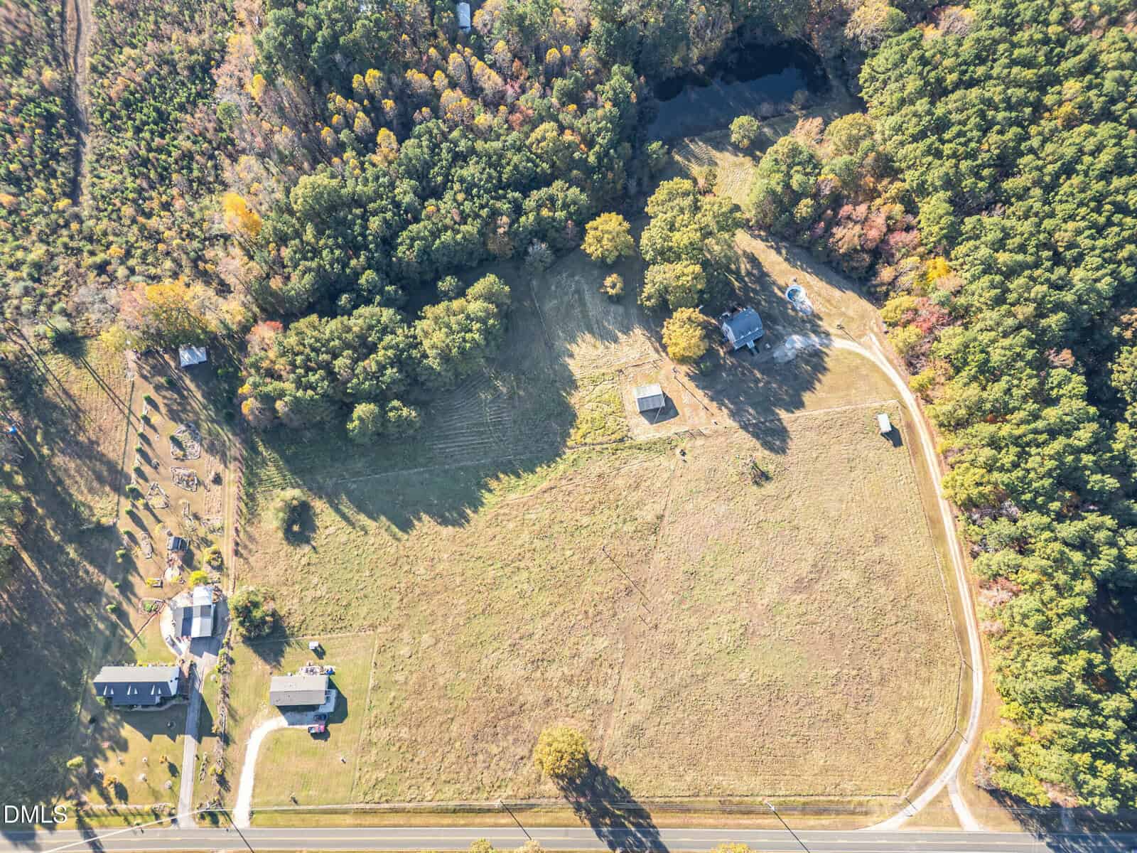Aerial view of a rural property with open grassy fields, scattered trees, a few small buildings, a house, a pond near wooded areas, and a curving driveway connecting to a main road at the bottom of the image.