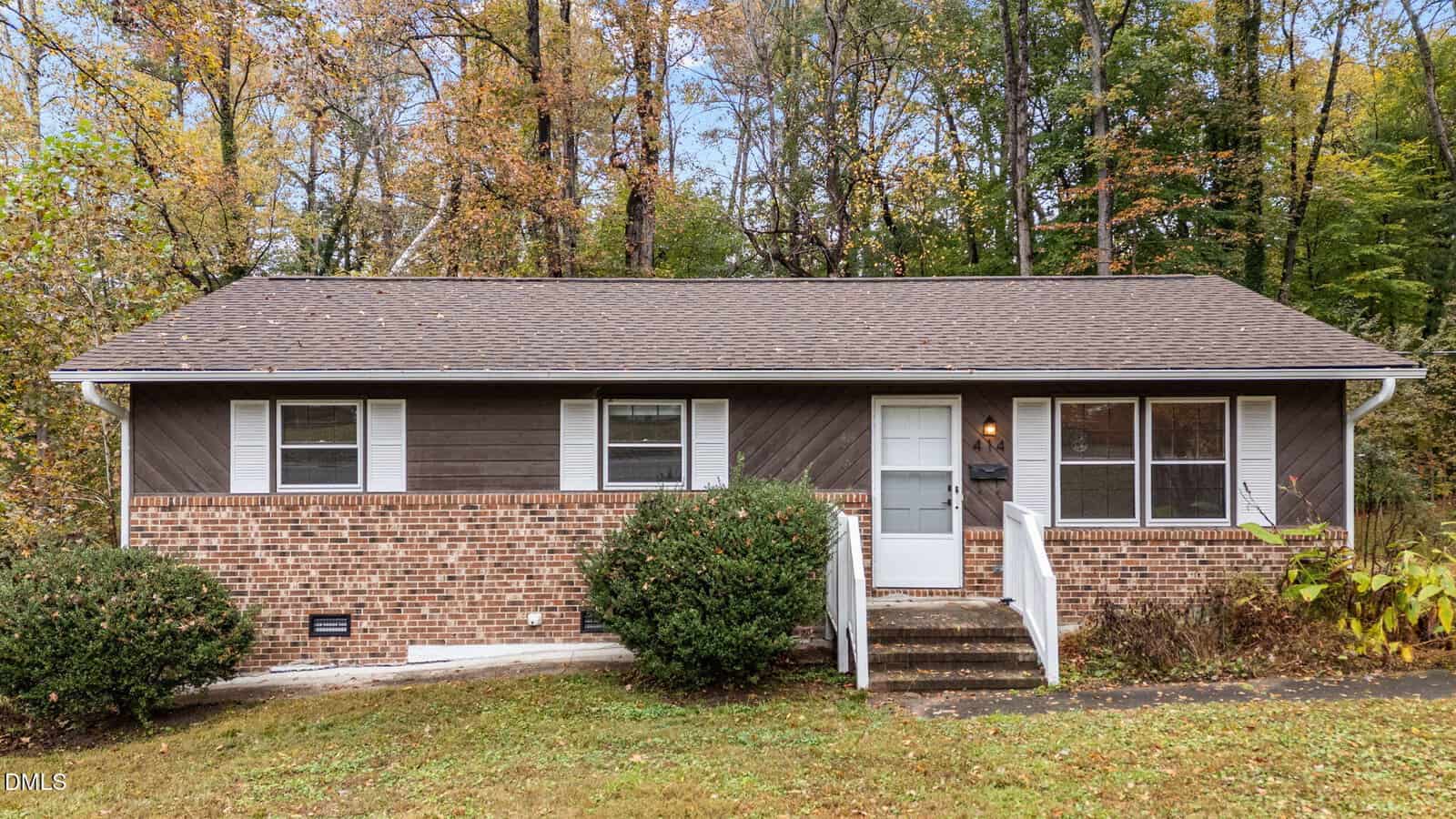 Single-story house with brown siding and partial brick facade, white trim, a shingled roof, and a central ramp with railings leading to the front door. Located at 414 W Noble Street in Louisburg, this home features two bushes in front and a wooded background with beautiful autumn foliage.