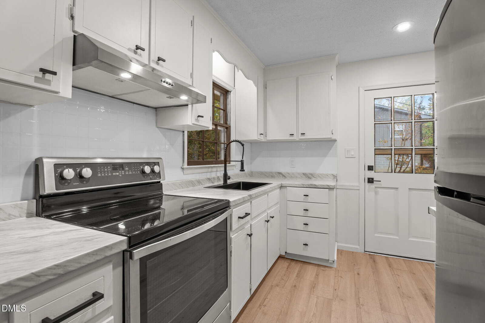 Modern kitchen with white cabinets, stainless steel appliances, a black faucet, marble countertops, a window above the sink, light wood flooring, and a white door with glass panes leading outside.